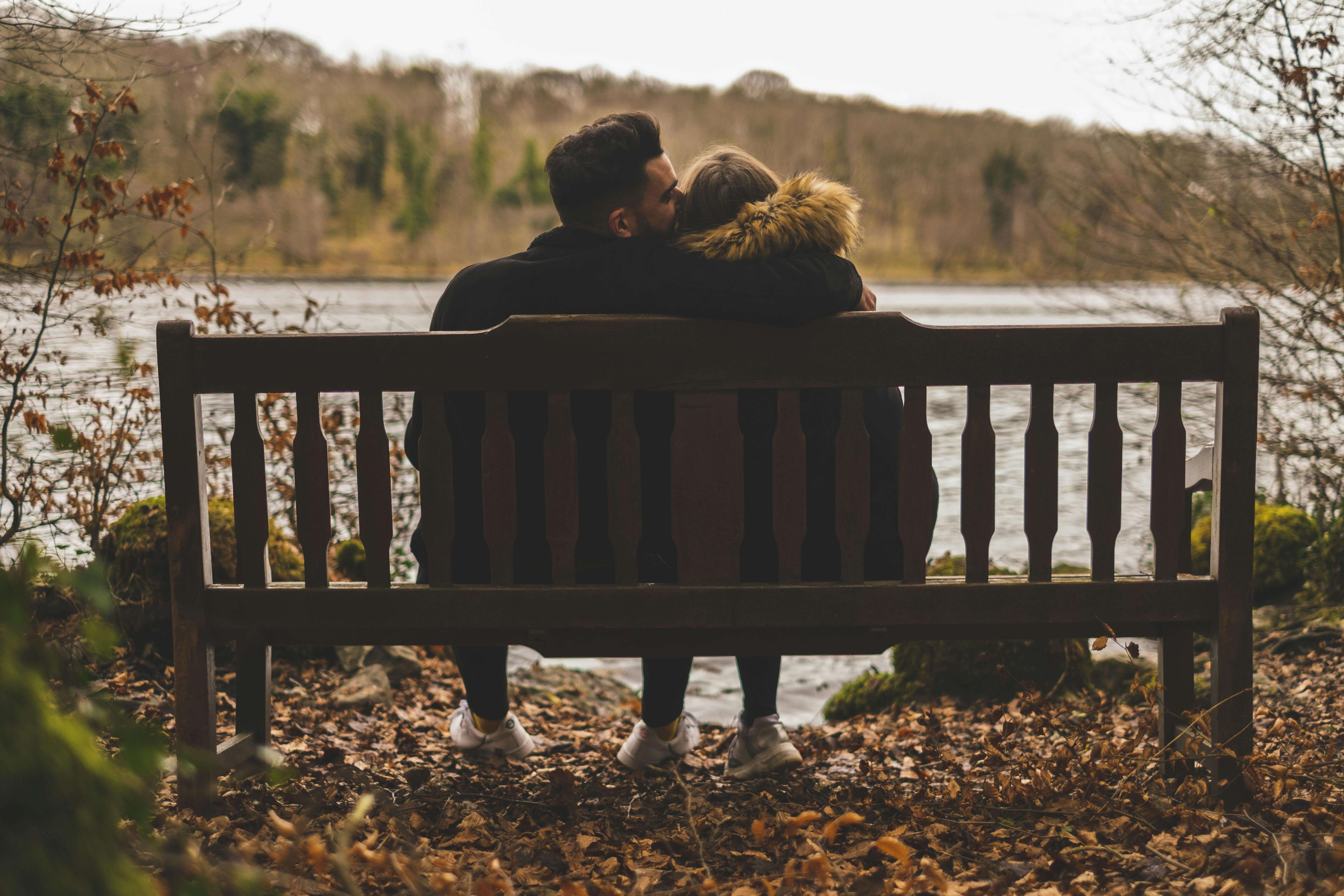 A couple shares a tender moment on a rustic bench by a serene lake. Captured in fall.