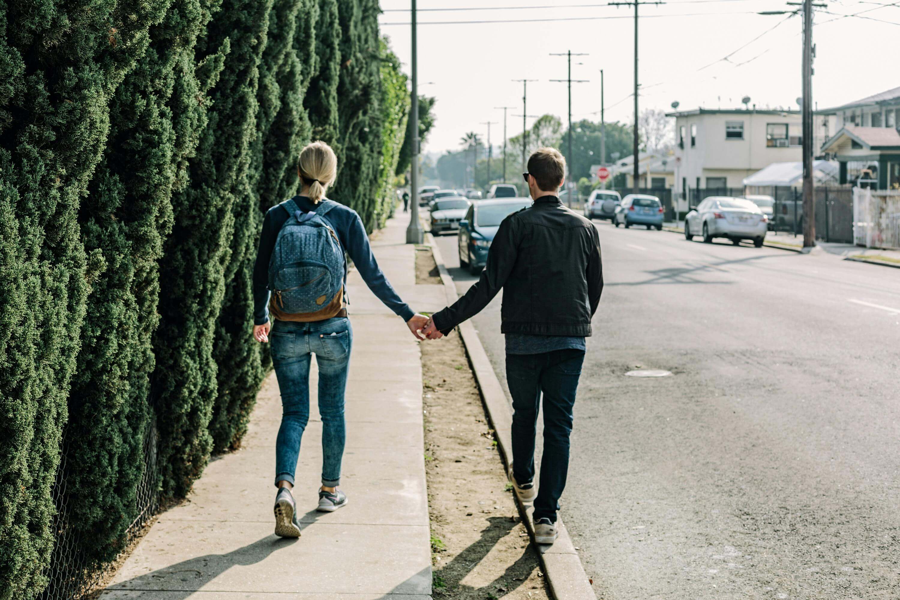 A couple walks hand in hand along a sunny sidewalk in an urban area, showcasing romance and companionship.
