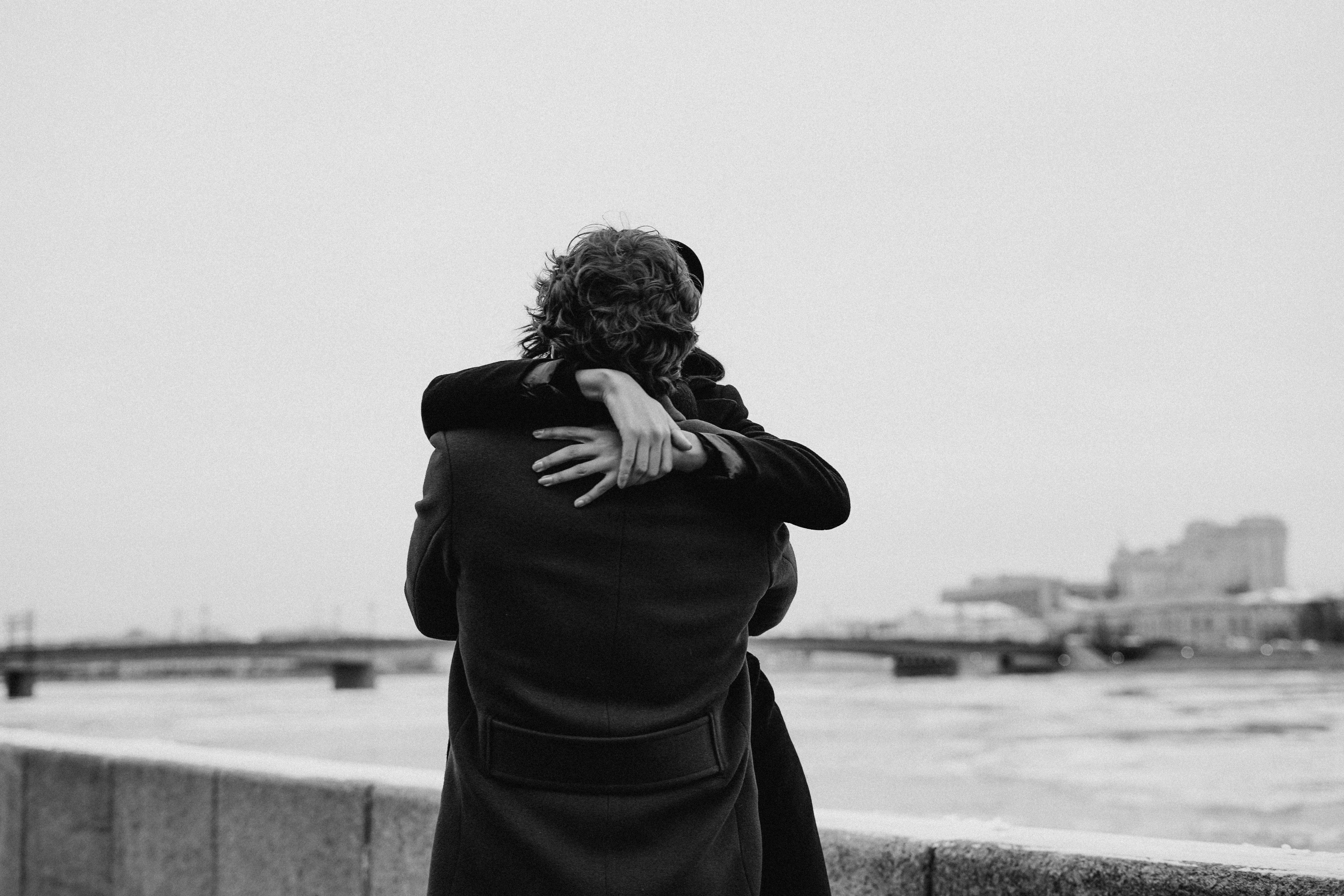 A couple embraces by the river in a monochrome winter setting, showcasing love and togetherness.
