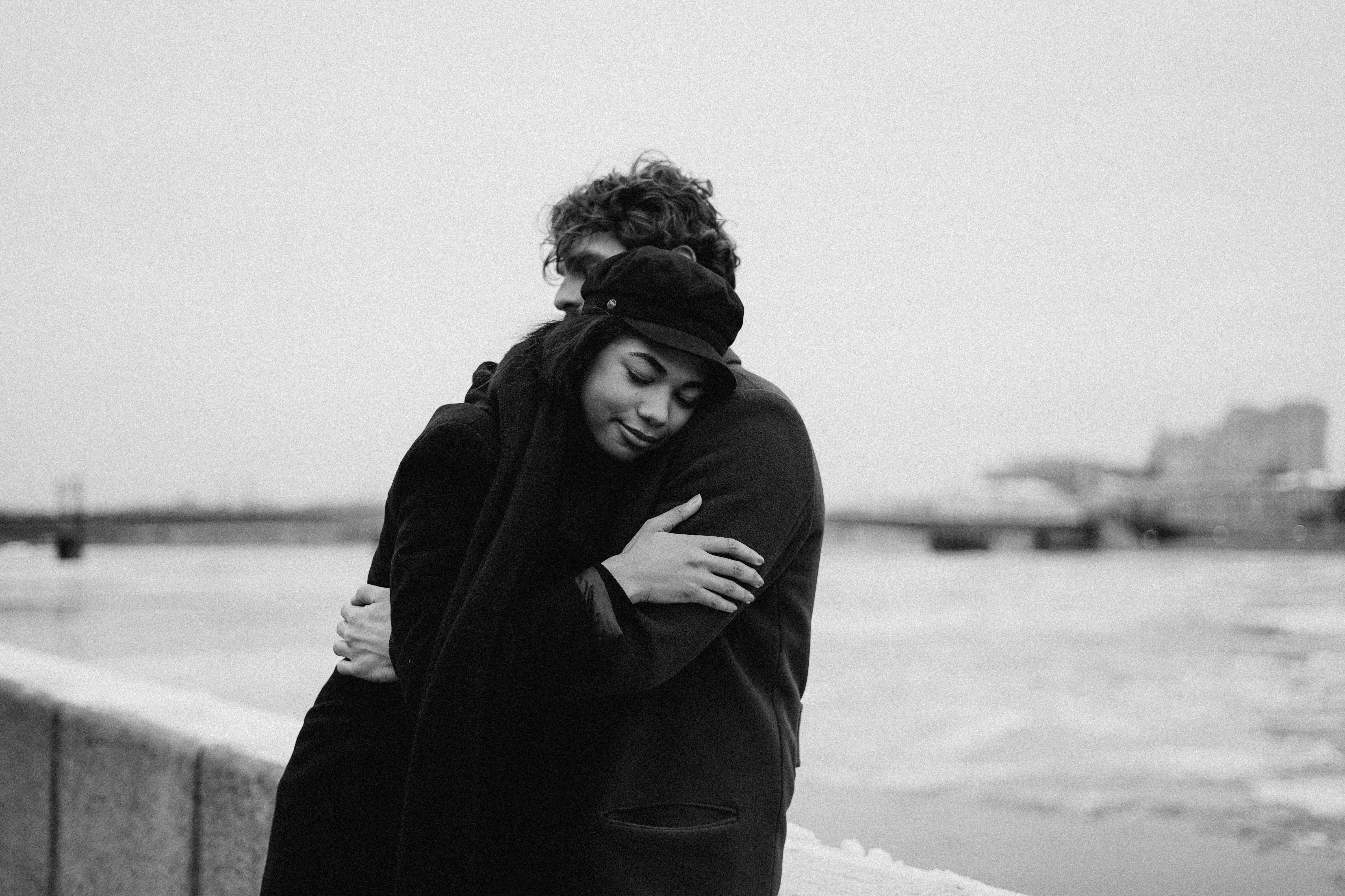 A loving couple shares a warm embrace outdoors by a river in a winter setting.