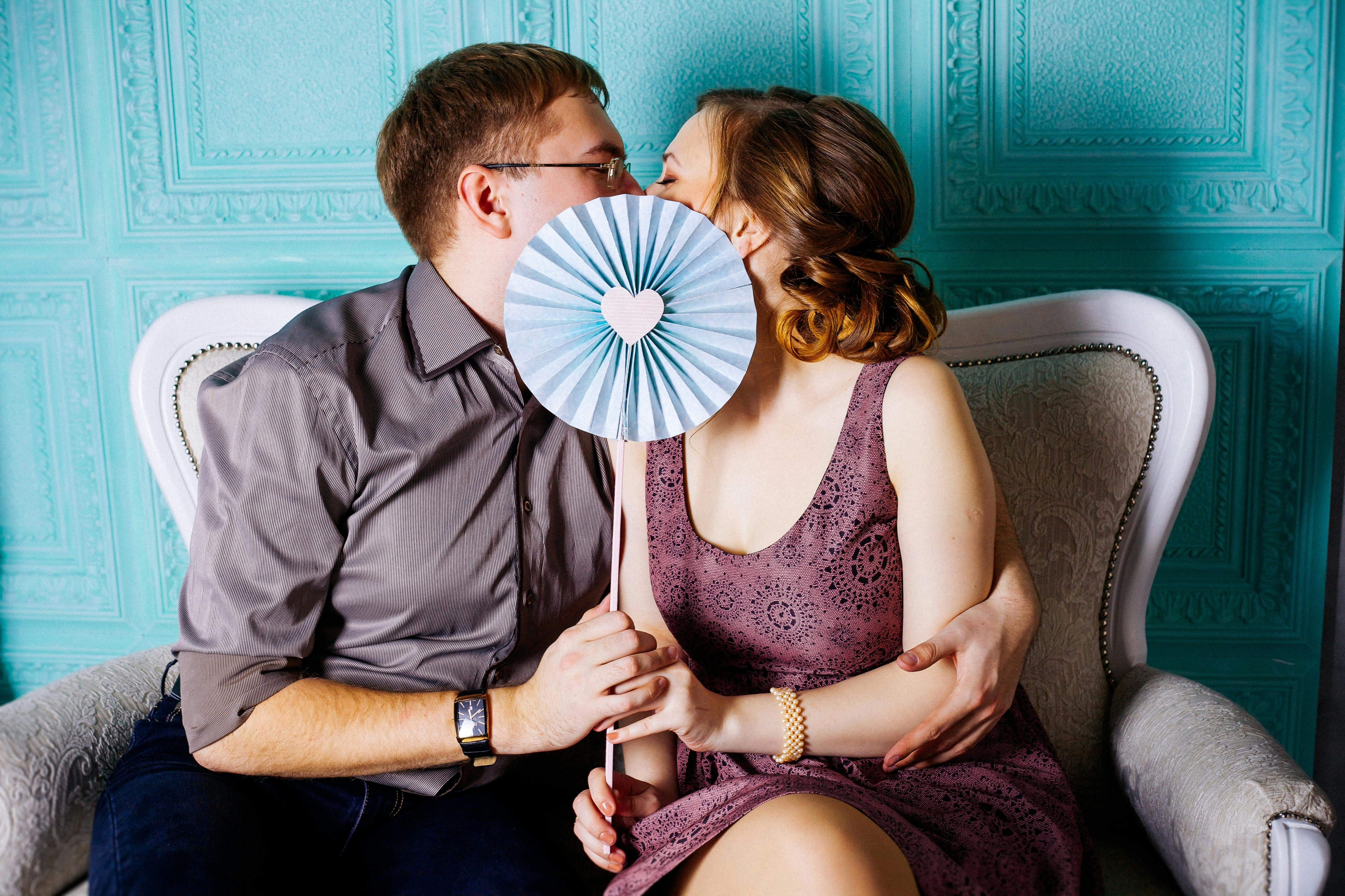 A couple sharing a kiss behind a heart-shaped decor, seated on an elegant chair indoors.