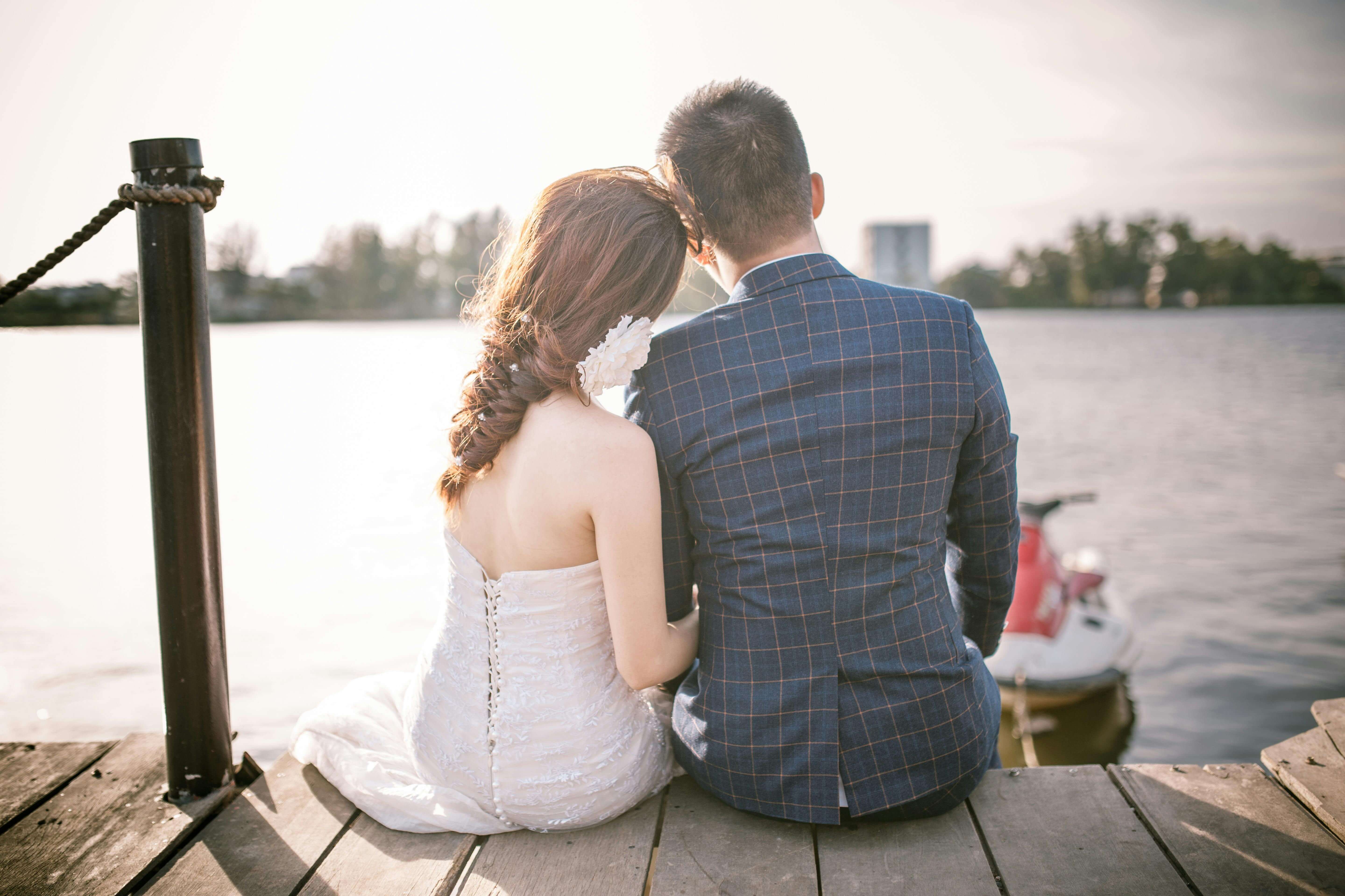 Bride and groom sitting on a dock by a serene lake, enjoying a romantic moment under the sun.