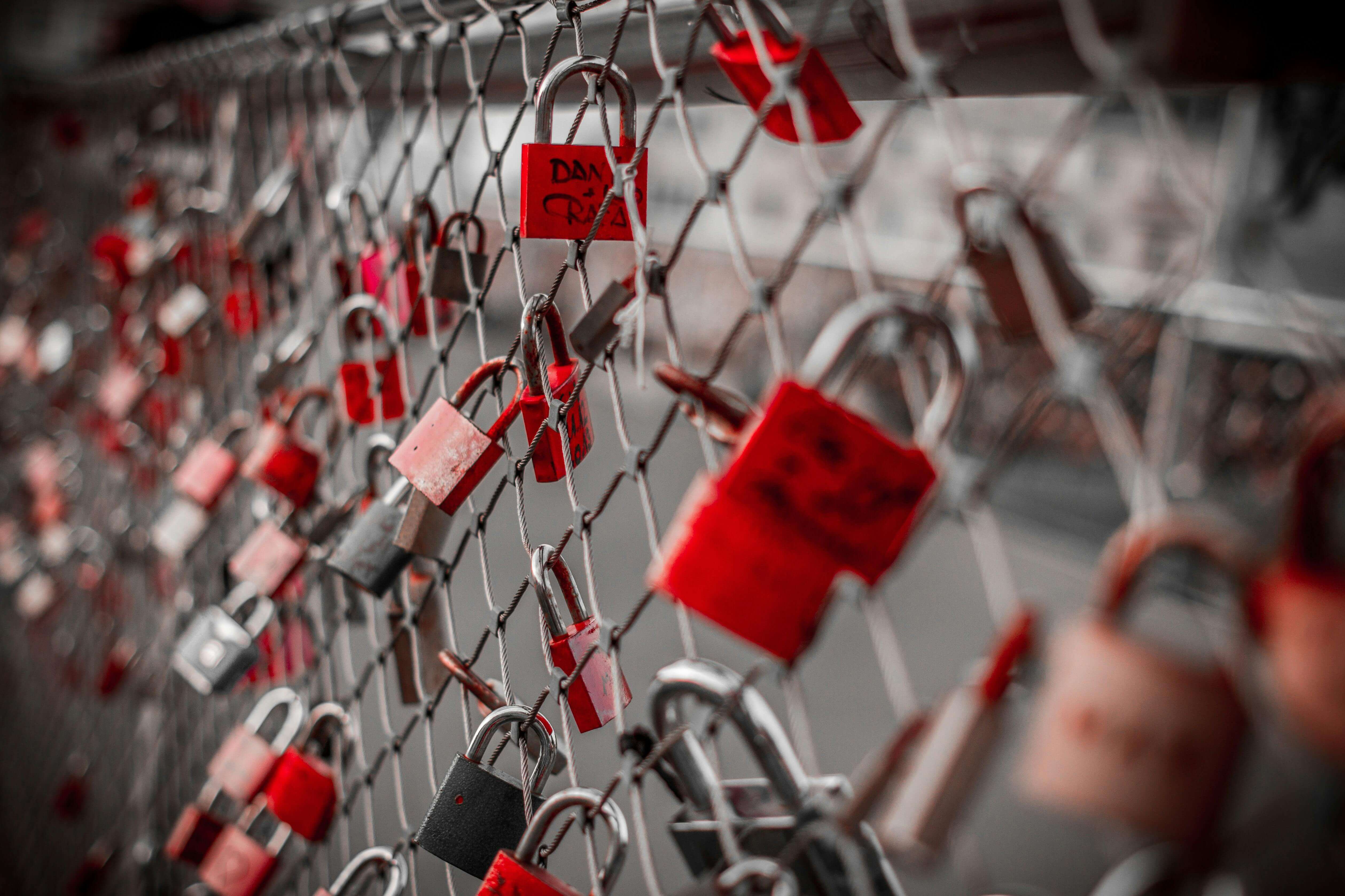 Red and silver padlocks symbolizing love attached to a chain link fence on a bridge.