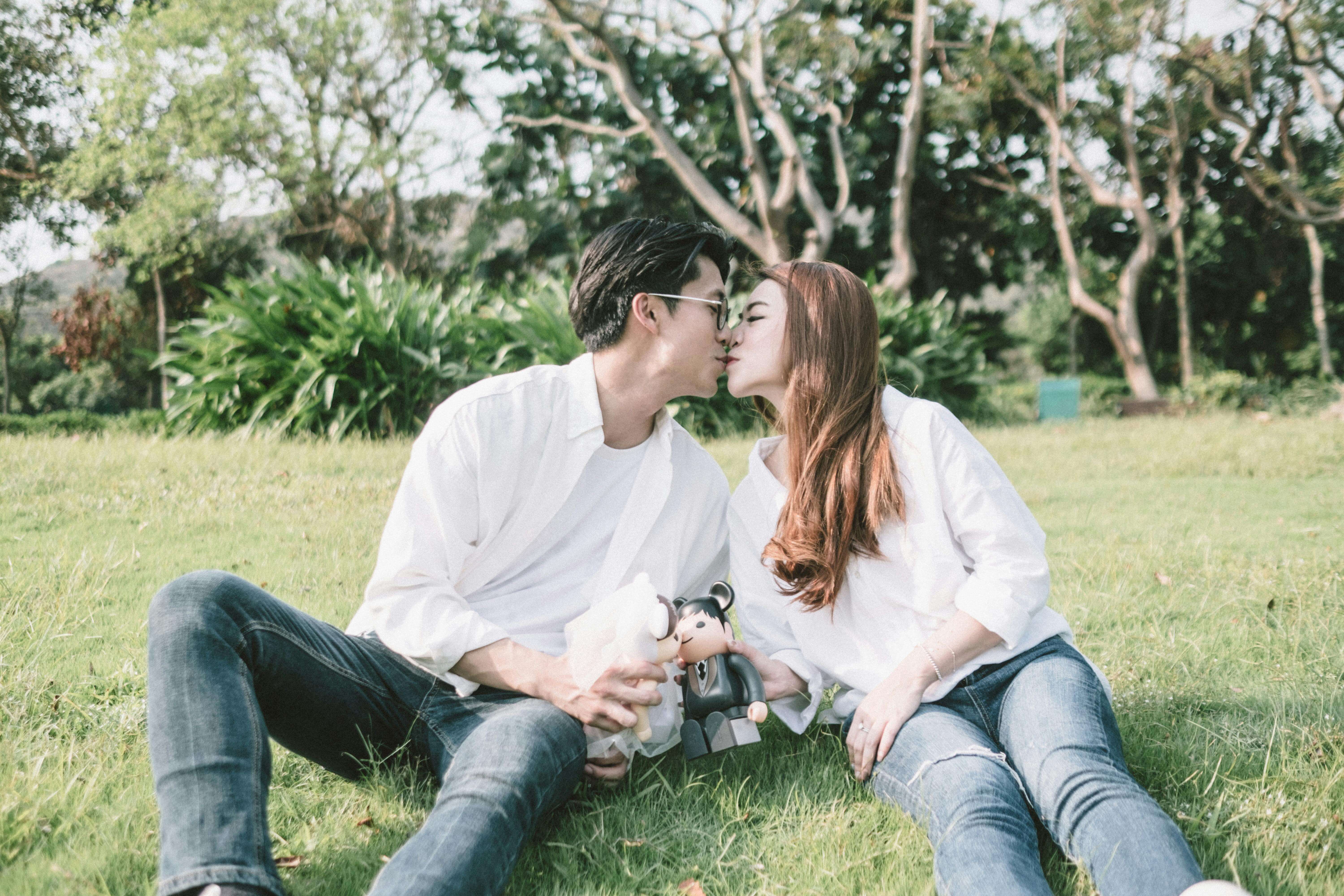 Young Asian male and female with figurines in hands sitting and kissing on grass in field in sunny summer day
