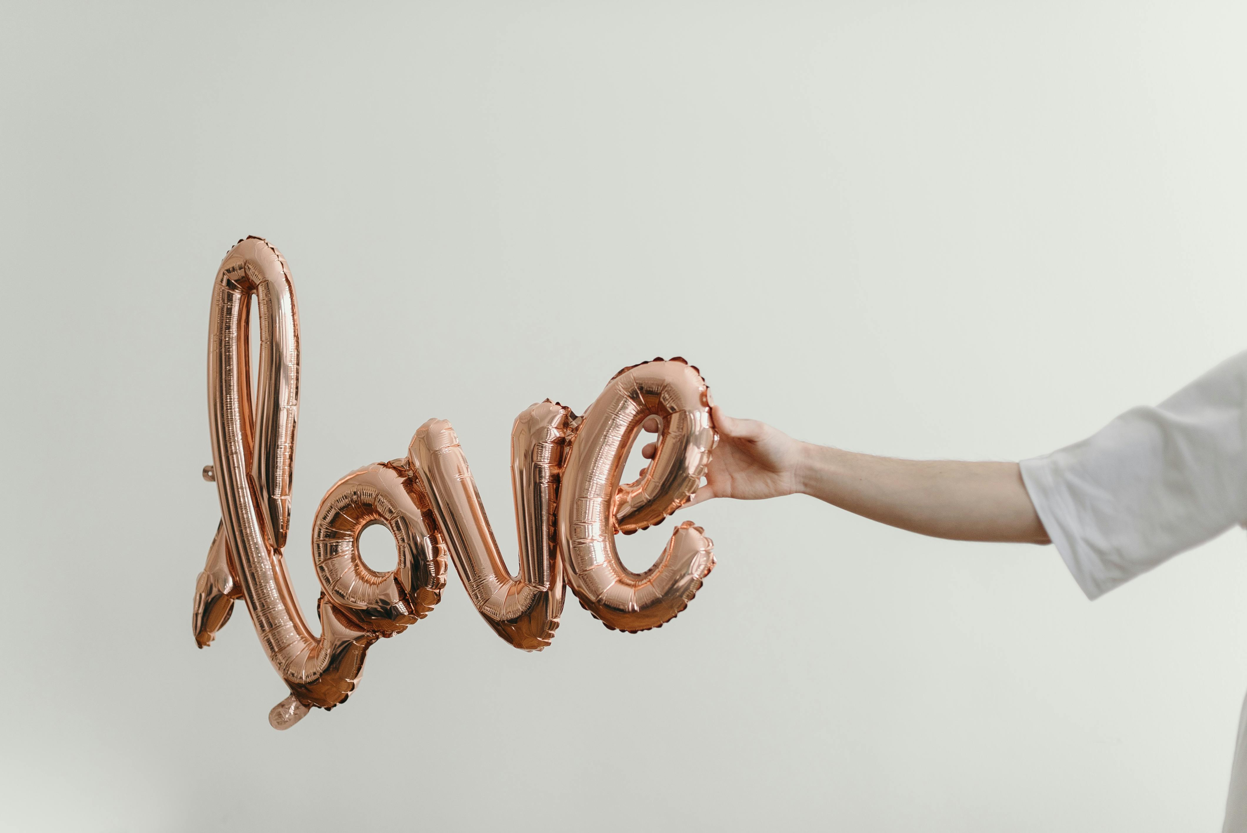 A faceless person holding a rose gold balloon spelling 'love' on a white background.