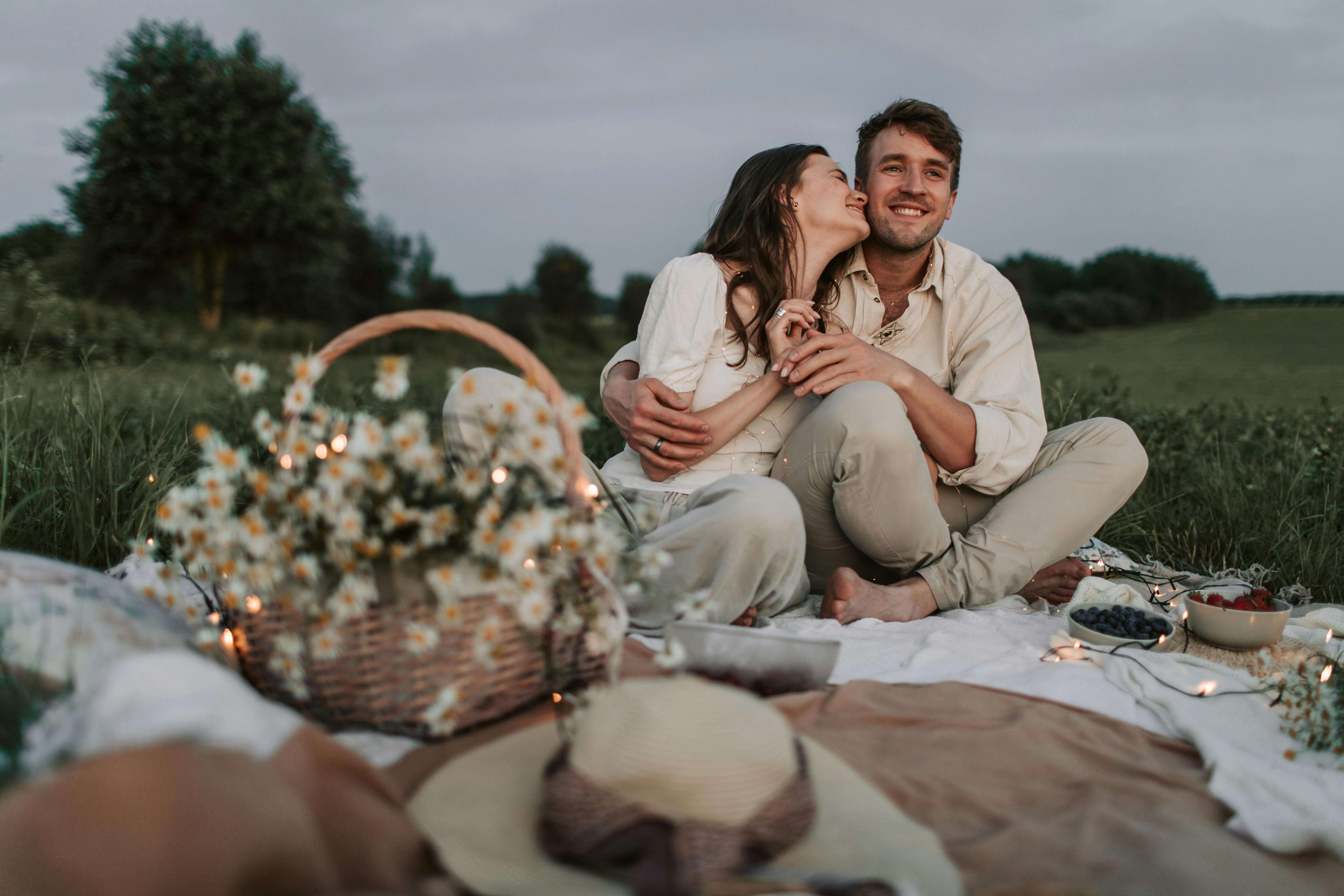 Couple sitting on a picnic blanket, embracing in a grassy field during a summer day.