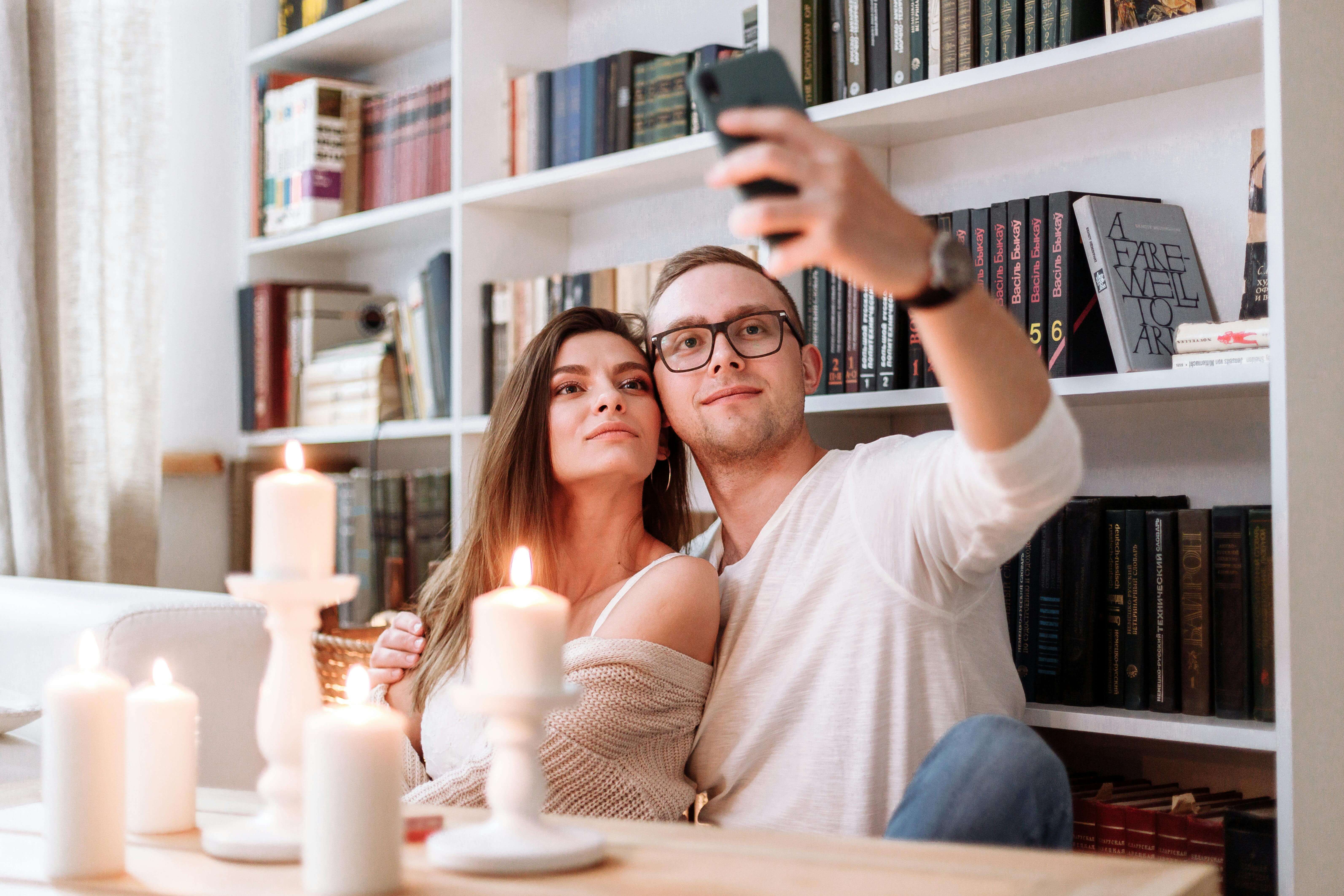 A happy couple taking a selfie while sitting in a cozy library, surrounded by books and candles.