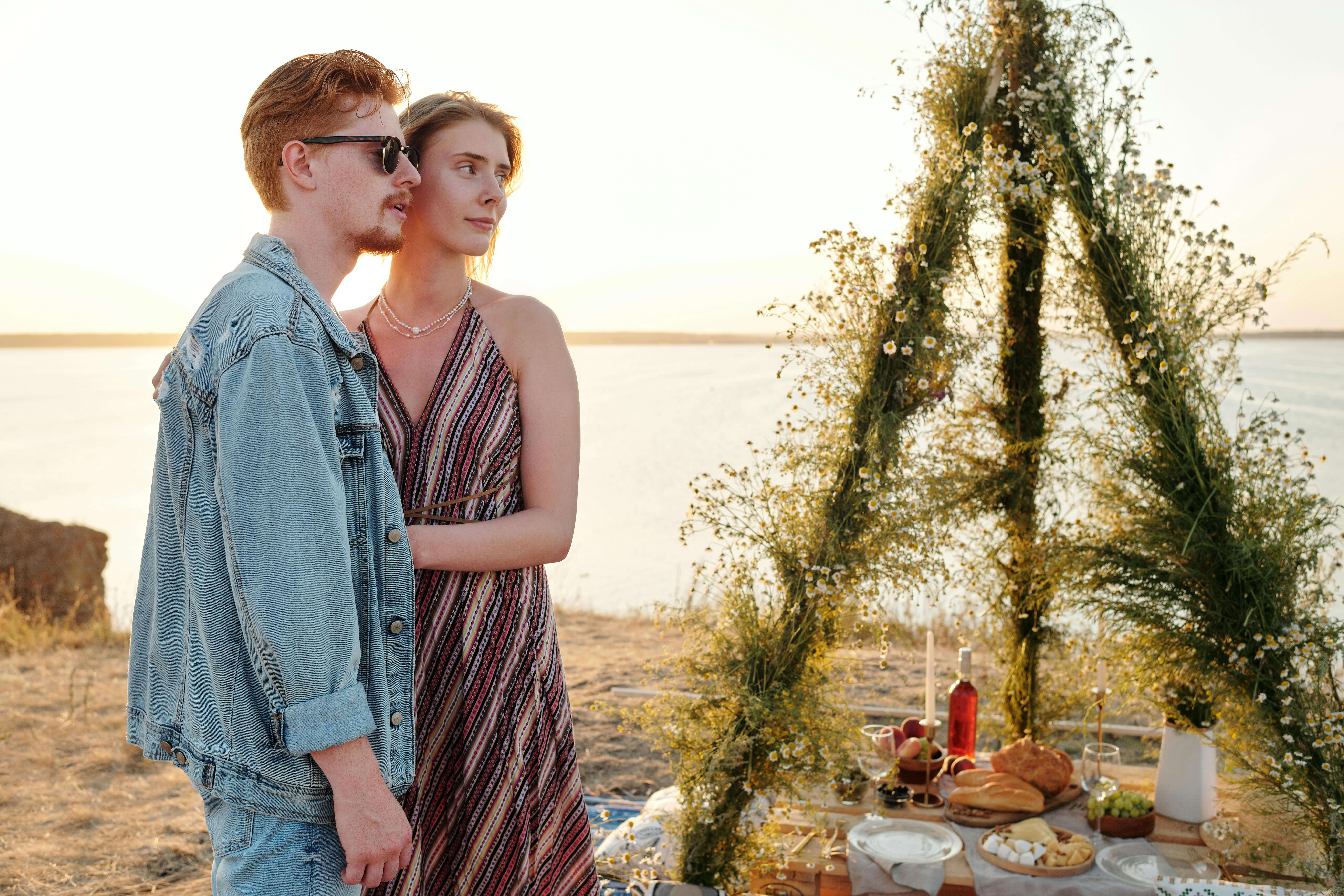 Couple embracing on a beachside picnic with food and floral decor at sunset.