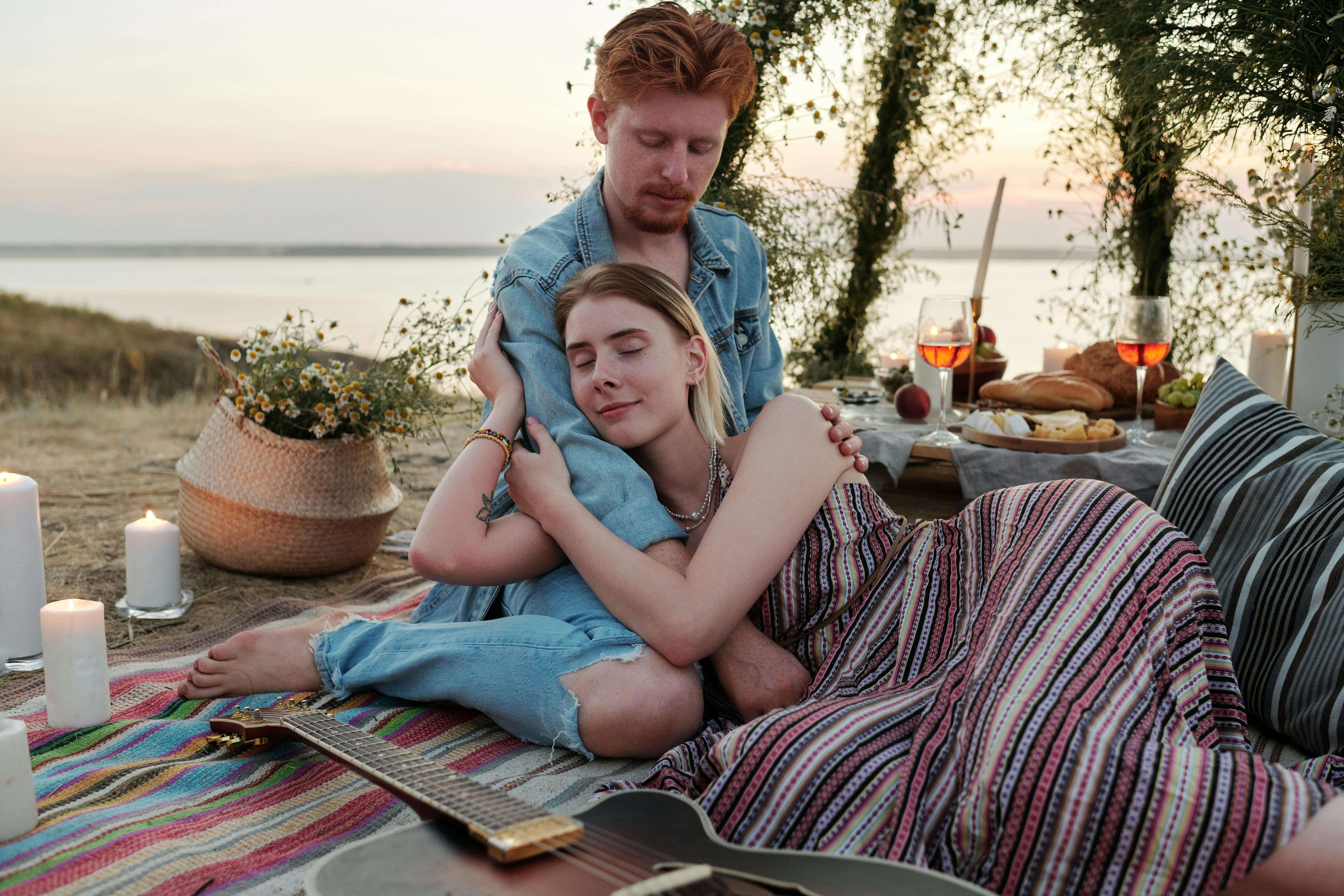 A couple enjoys a cozy lakeside picnic with candles and wine at sunset.