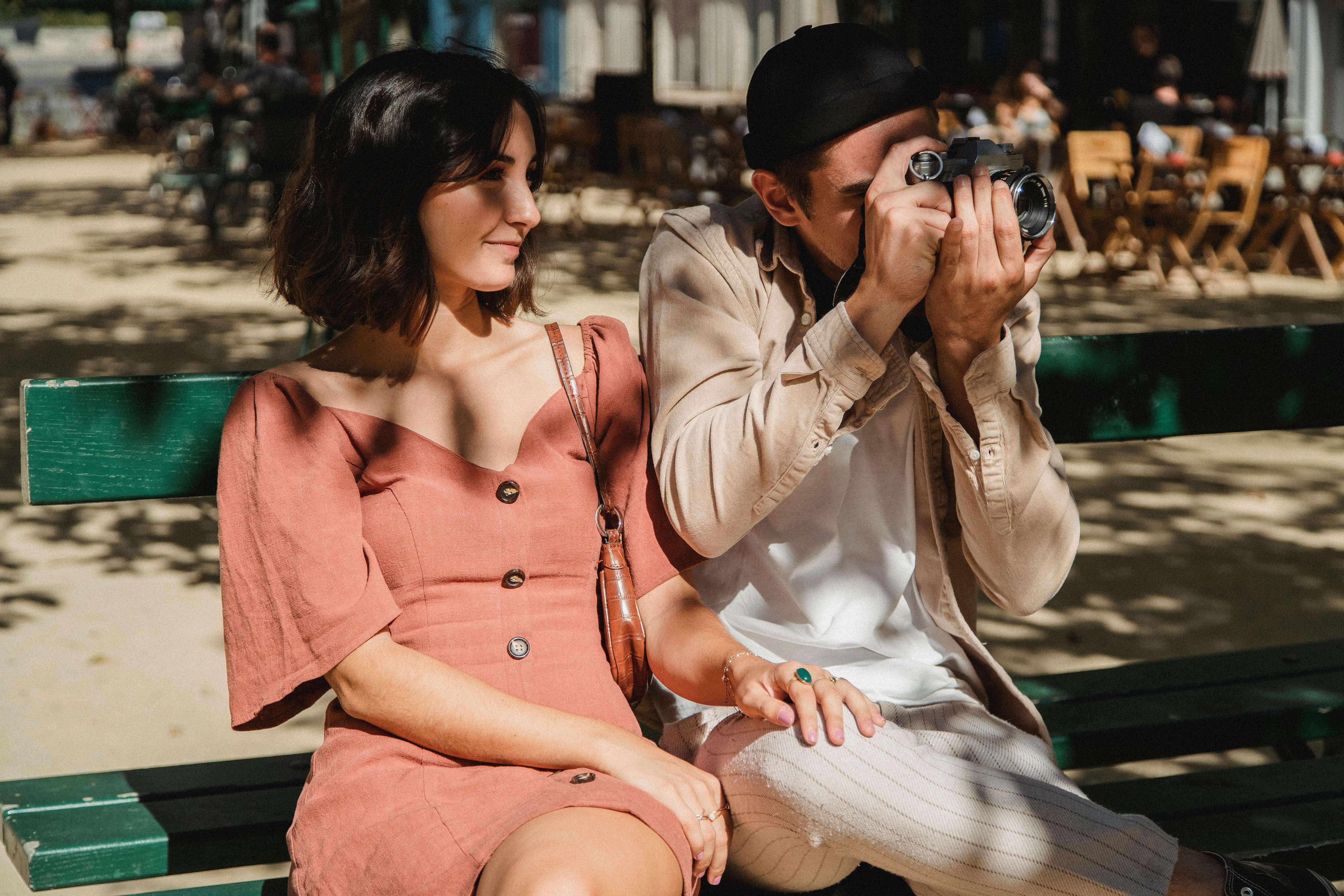 Couple enjoying a sunny day on a park bench while taking photos, capturing moments.