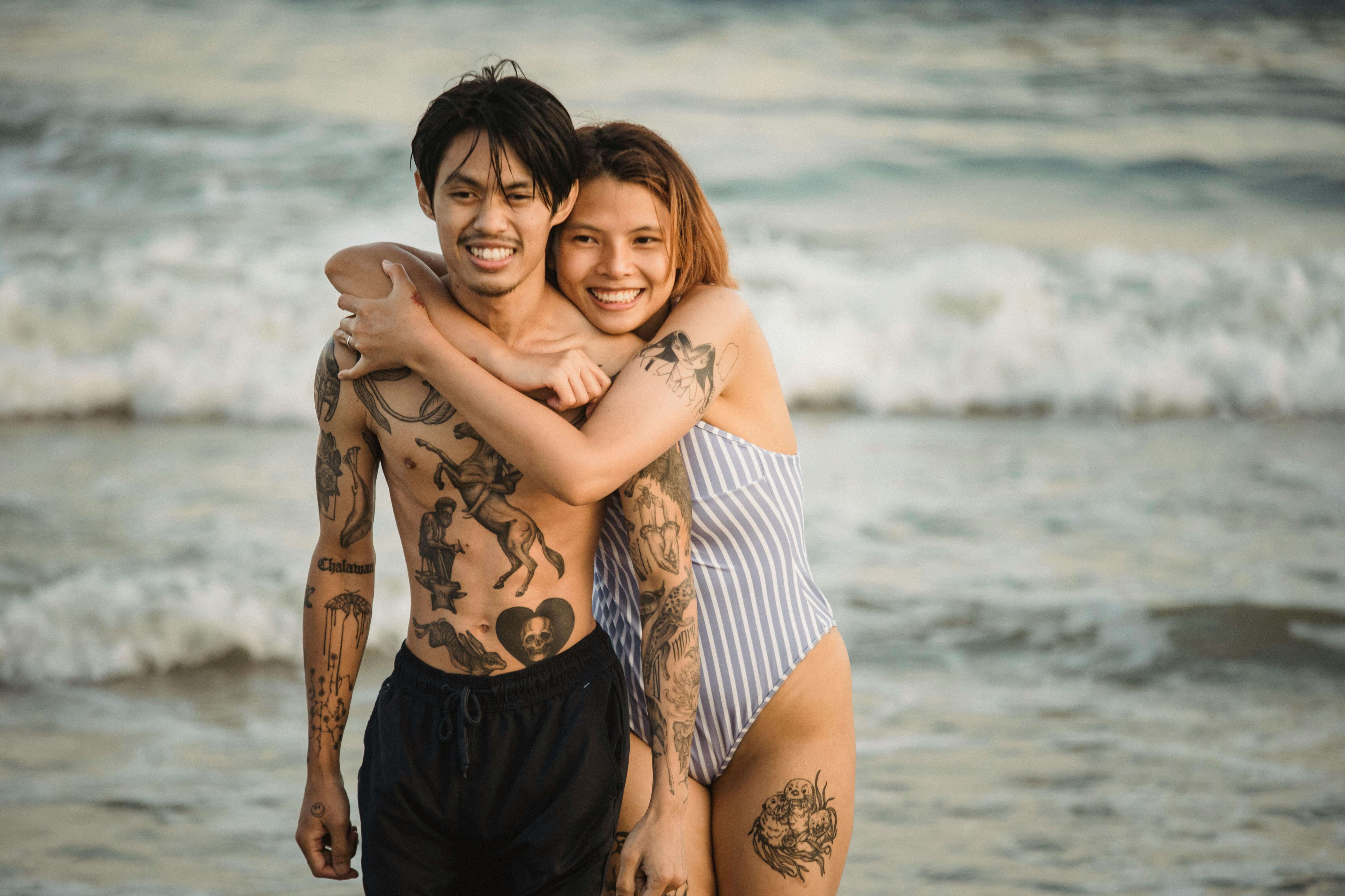 A joyful couple embracing by the seaside, enjoying a summer day with tattoos displayed.