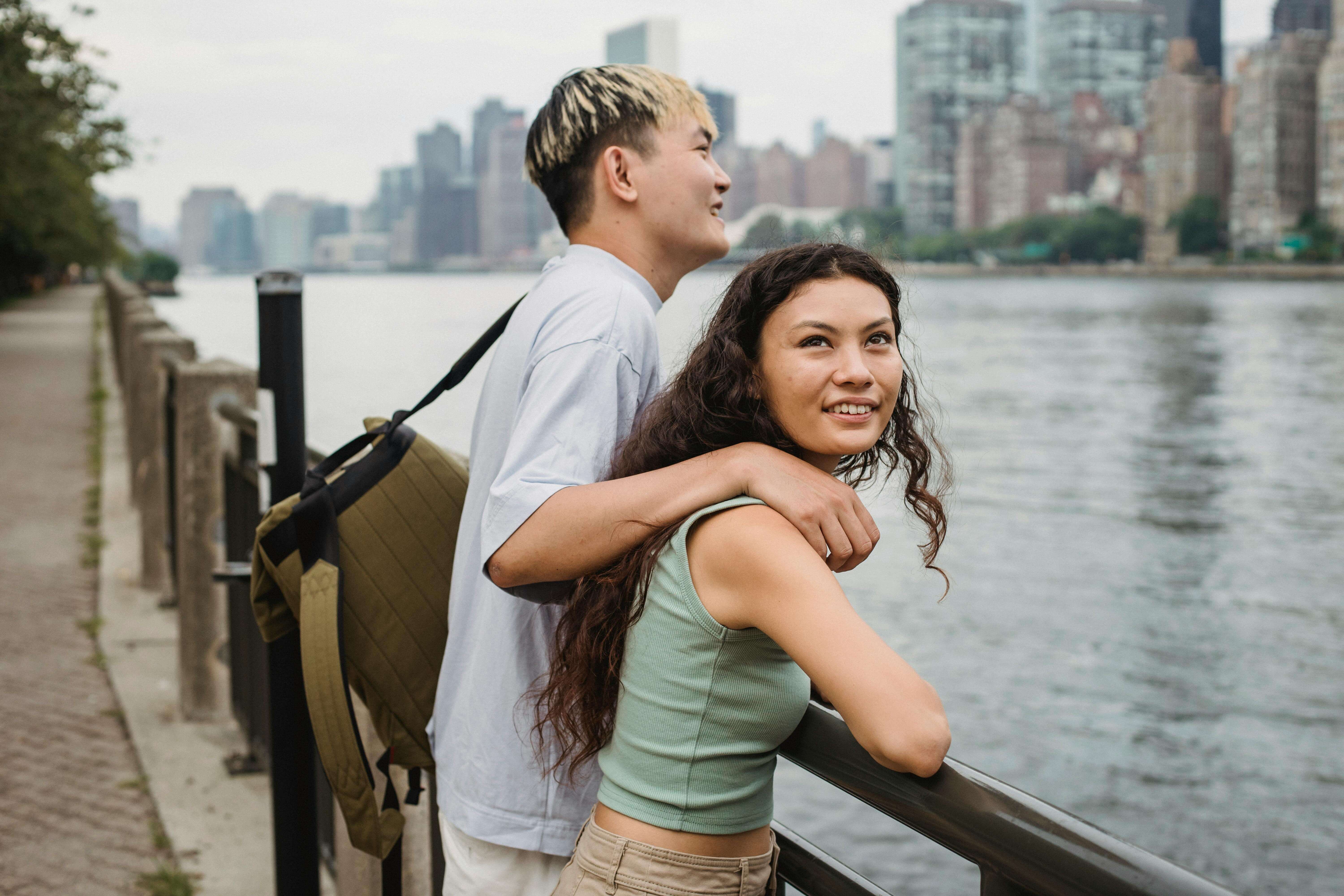 Side view of cheerful young diverse couple in casual clothes cuddling and smiling while standing on river embankment and sightseeing city