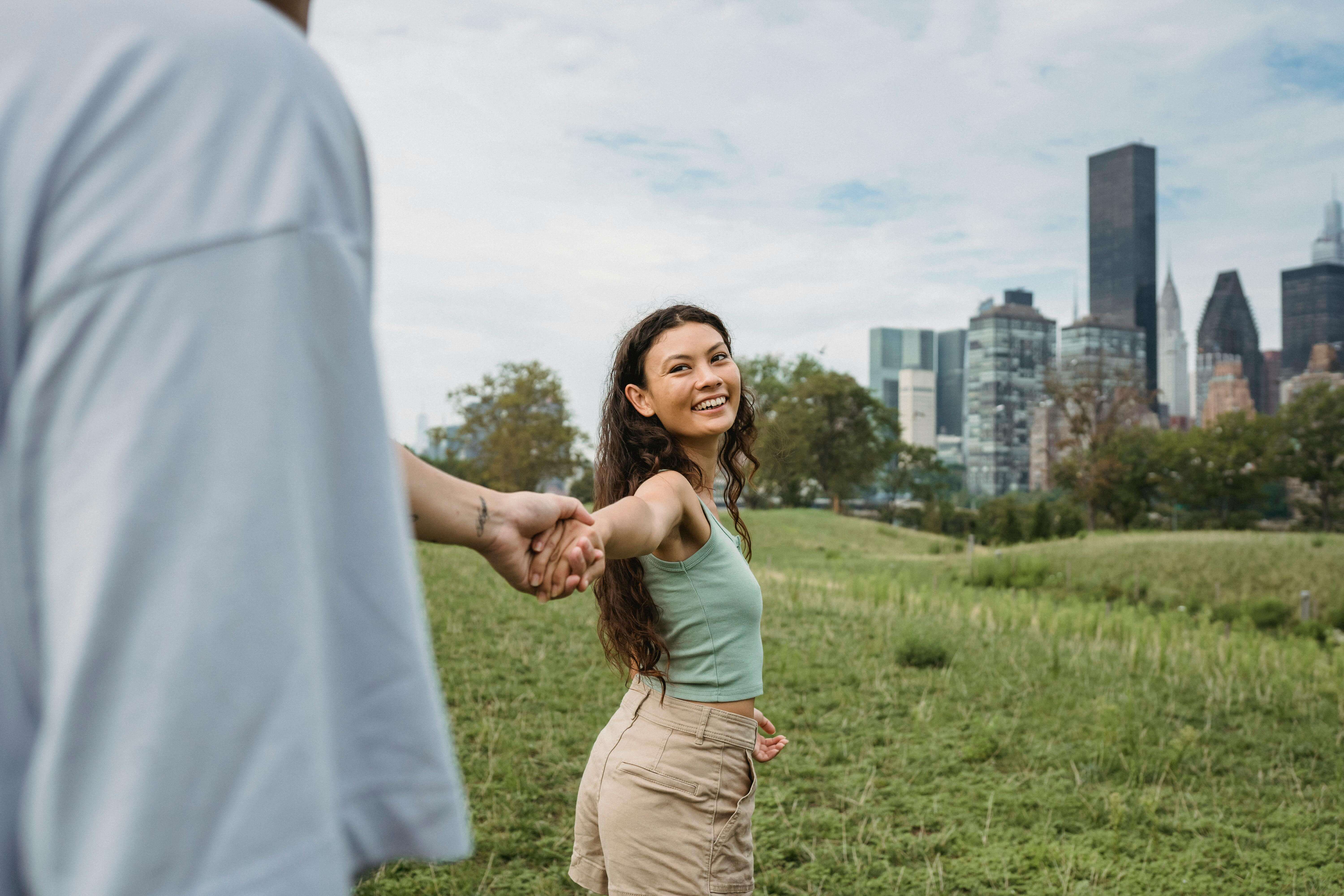 Crop anonymous man holding hand and following happy young ethnic girlfriend while spending time together in green park in New York