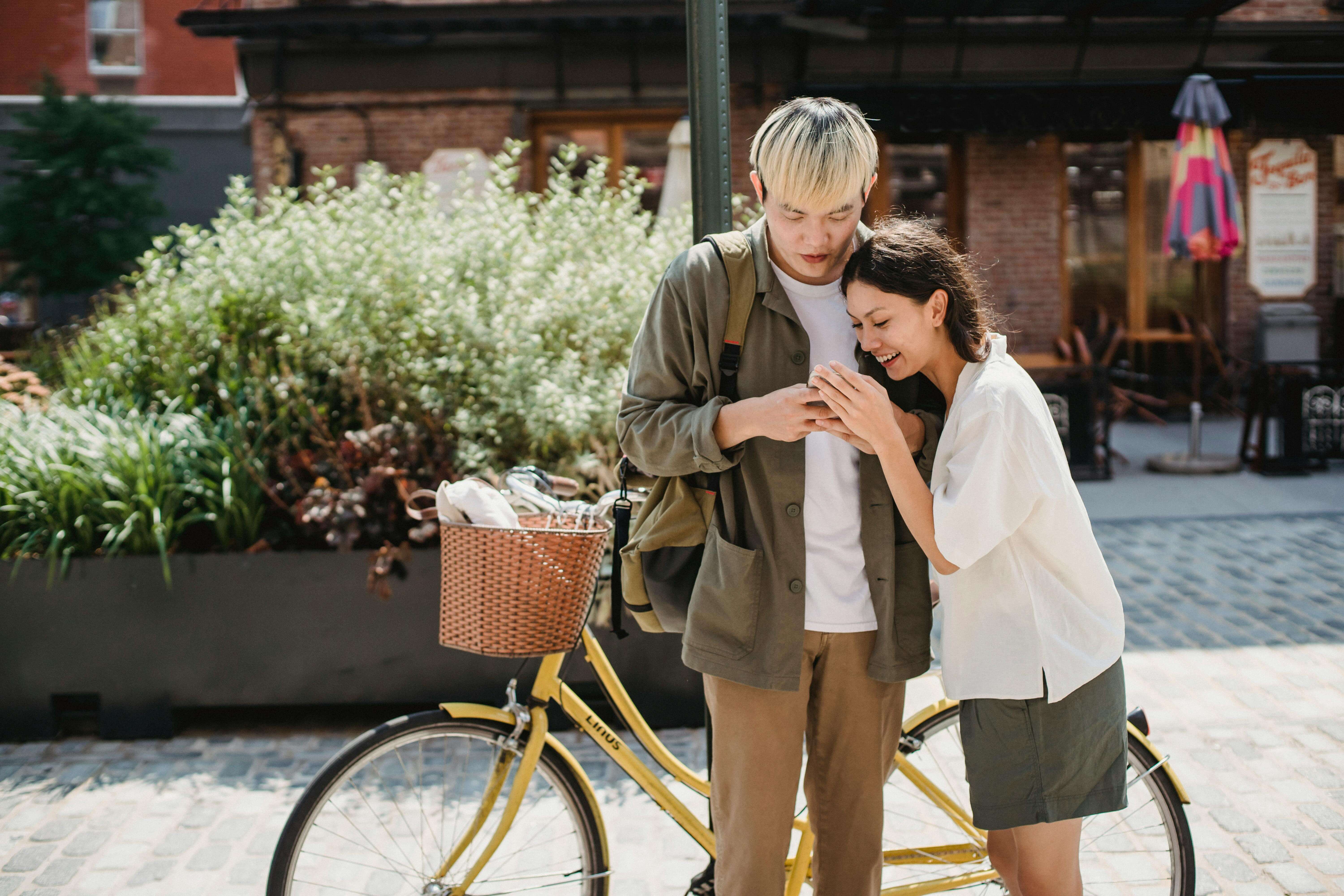Smiling young ethnic couple in trendy clothes watching photos on mobile phone while standing near stylish bicycle on street