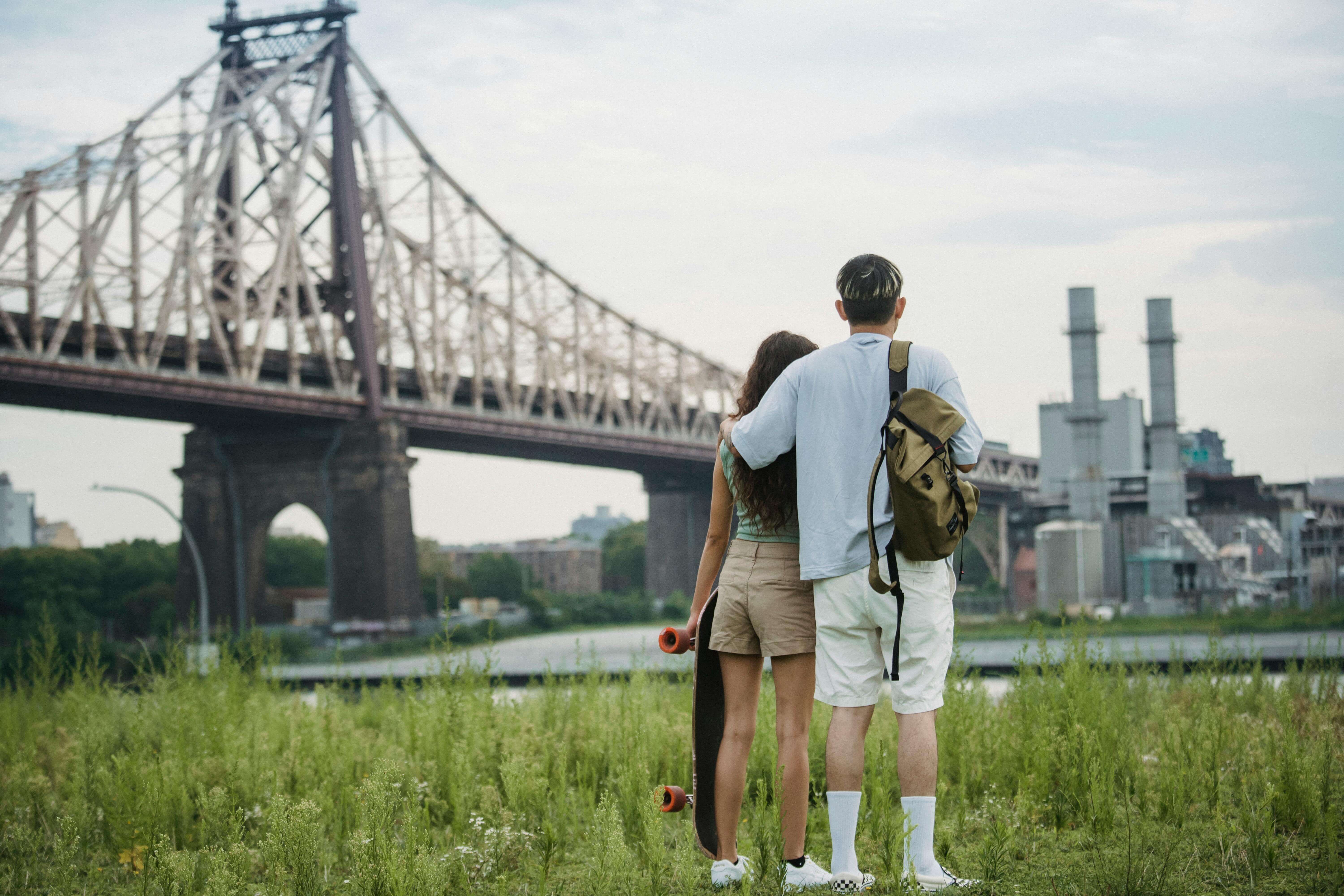 Back view of unrecognizable young male tourist in casual clothes with backpack cuddling girlfriend with skateboard in hand while standing on grassy ground near Brooklyn Bridge and admiring city