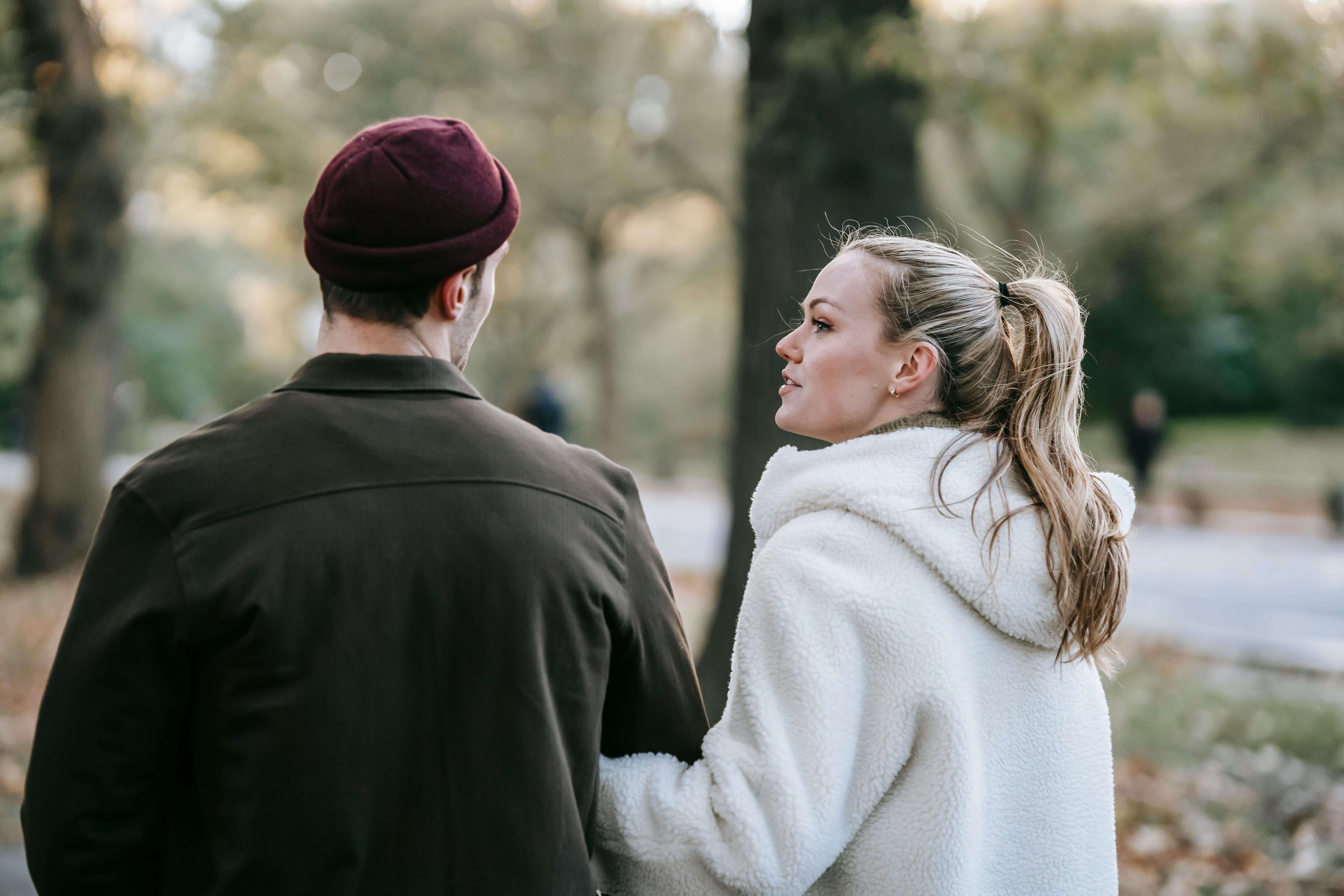 Back view of young stylish lady with blond hair in stylish warm outerwear holding hand of anonymous boyfriend while strolling together in autumn park