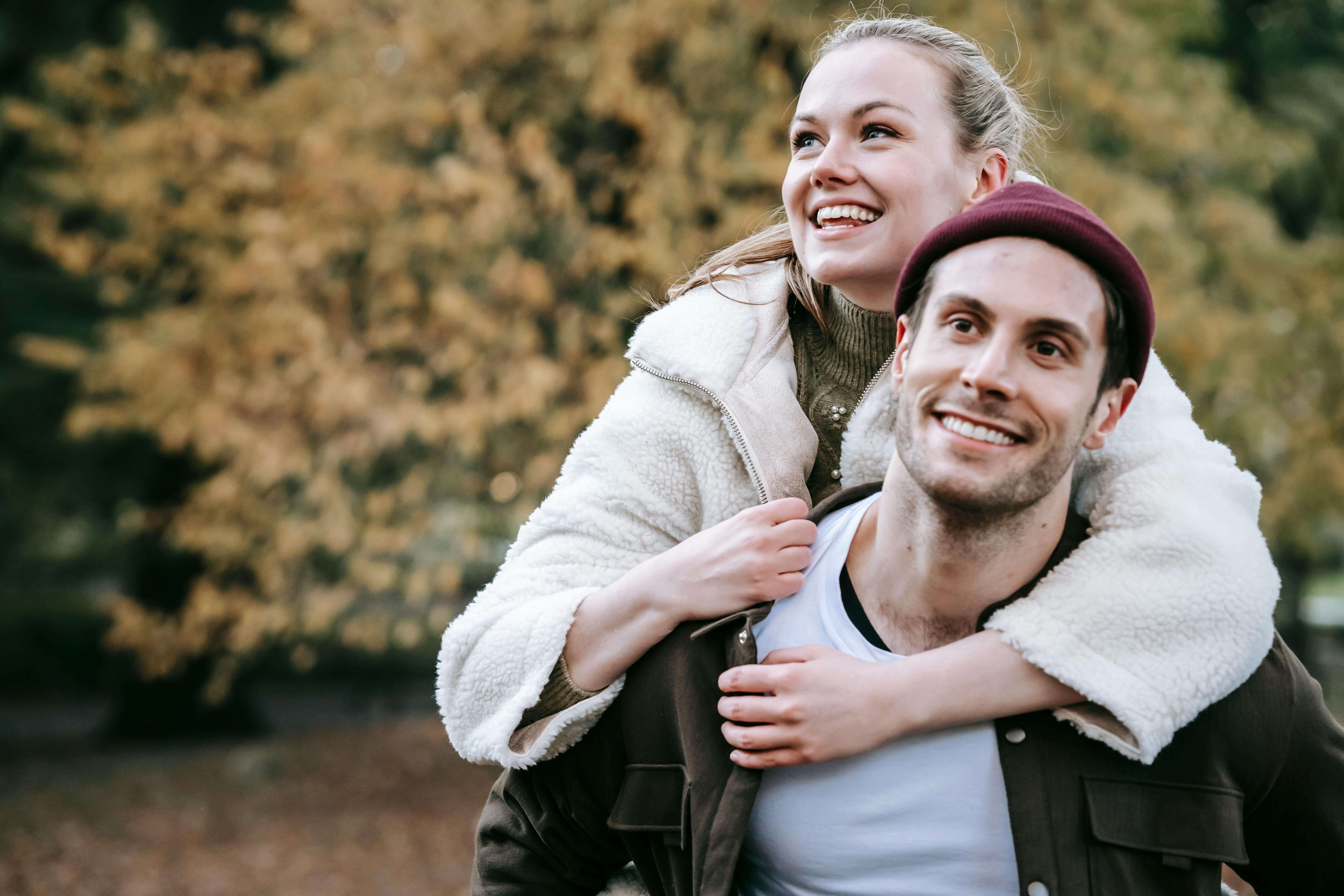 Positive young bearded boyfriend giving piggyback ride to smiling girlfriend on blurred background of trees with yellow foliage