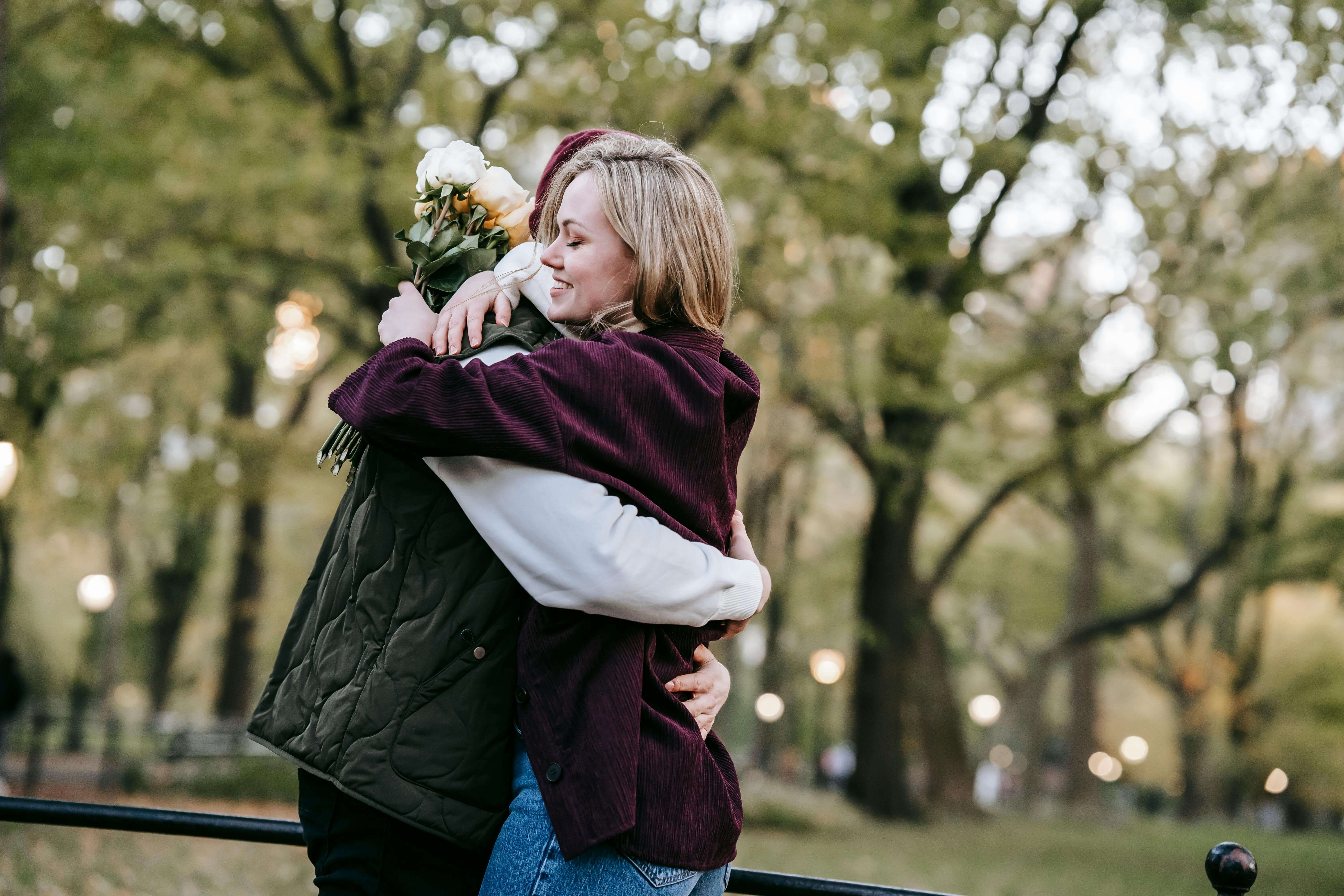 Side view of happy young couple in casual outfits with bouquet of flowers embracing in park