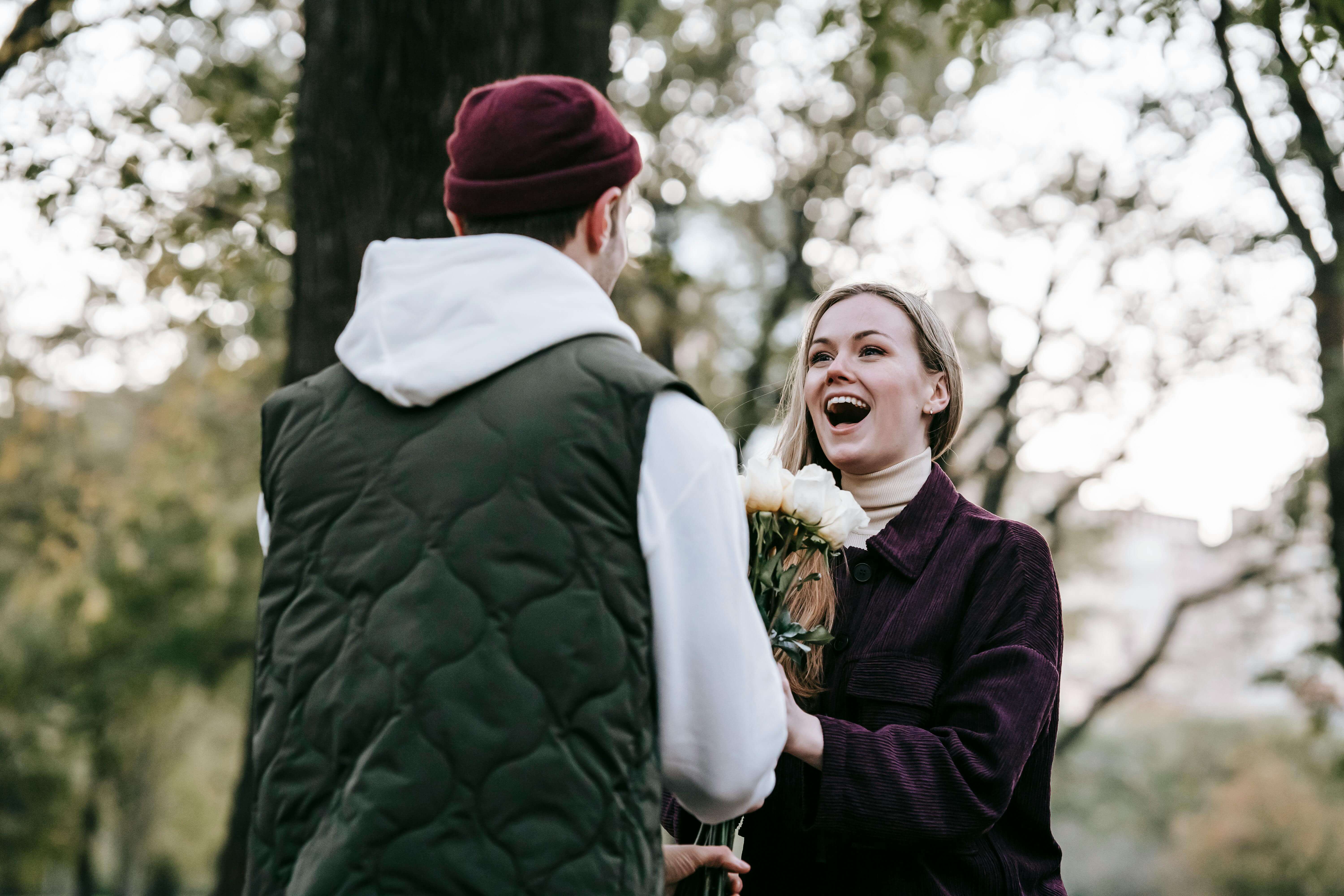 Loving boyfriend in hat giving bunch of roses as gift to happy girlfriend with long hair in casual warm clothes in park