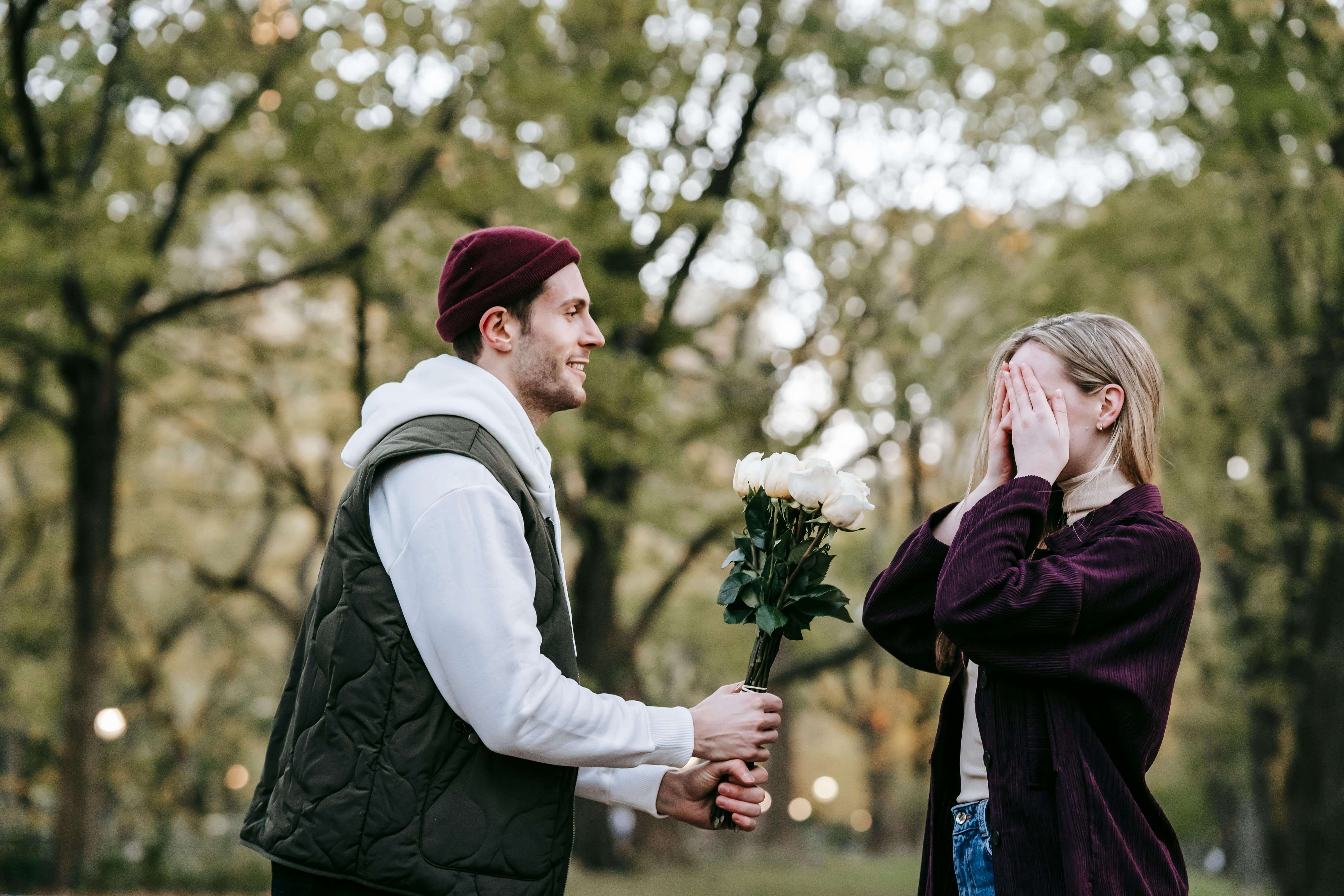 Side view of young loving man giving white roses as surprise present to girlfriend covering eyes with hands during date in park