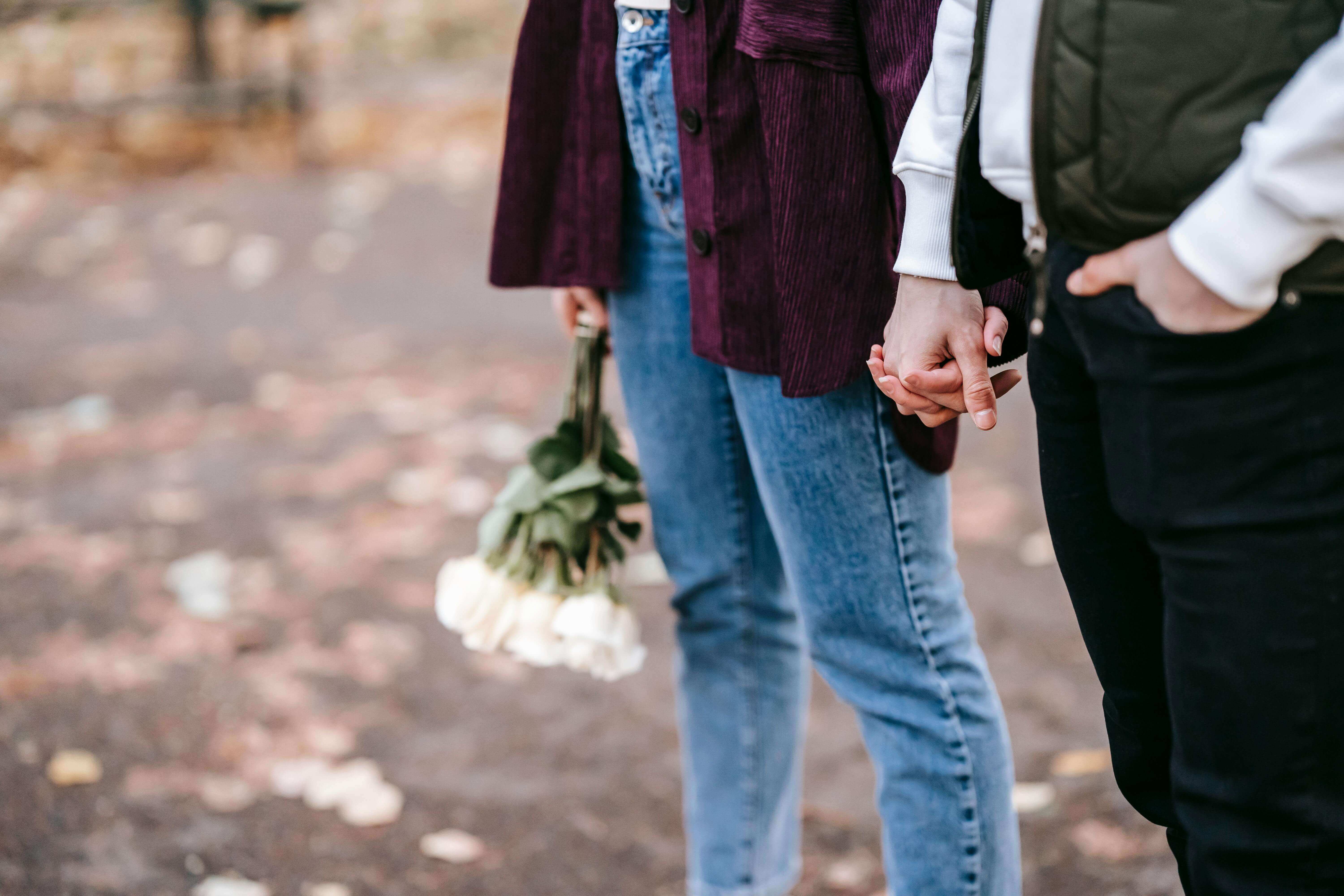 Crop anonymous couple with fresh white roses holding hands on blurred background of park