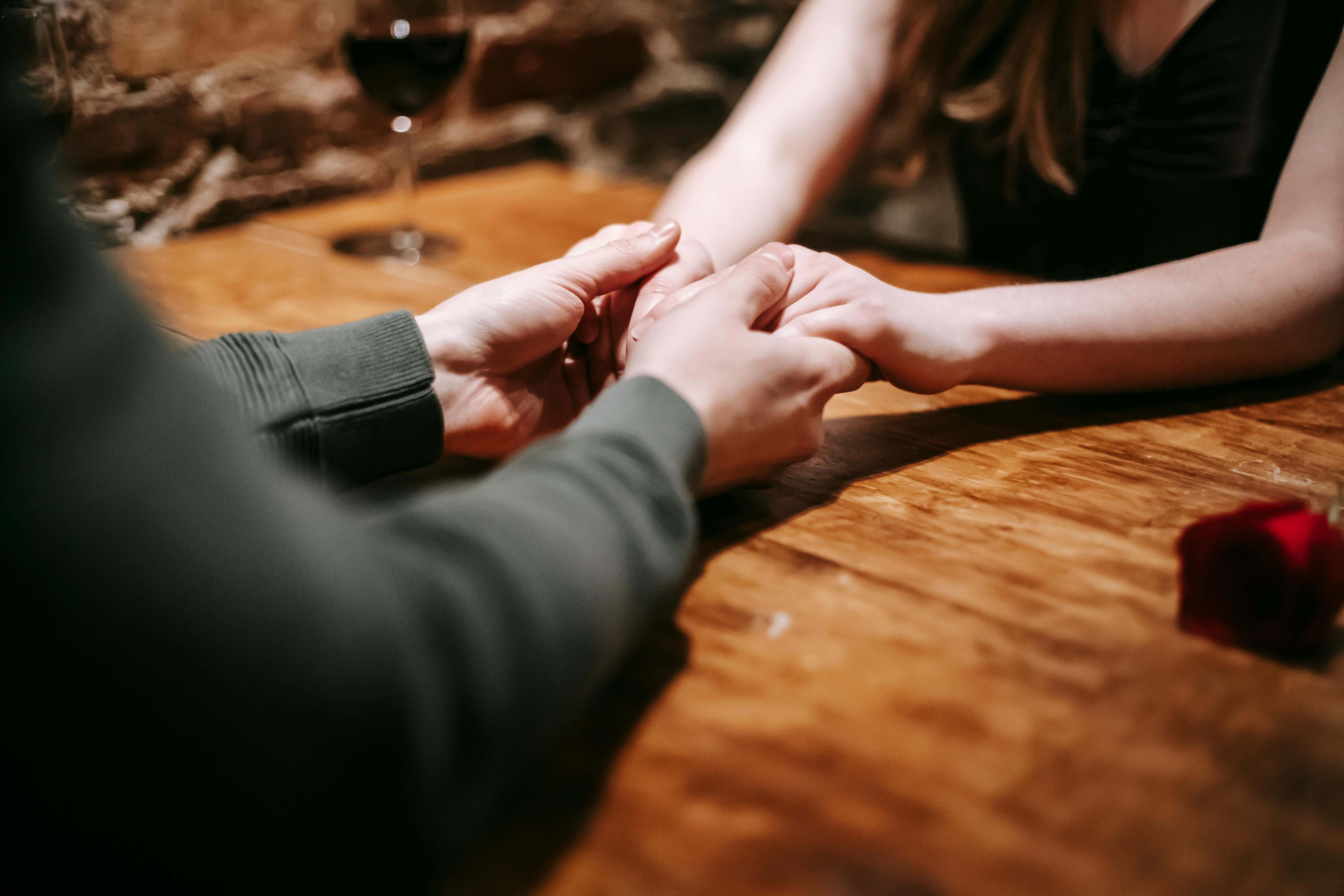 Crop anonymous couple sitting in light cafeteria at wooden table with glass with wine and red rose near brick wall and having romantic dinner while holding hands gently