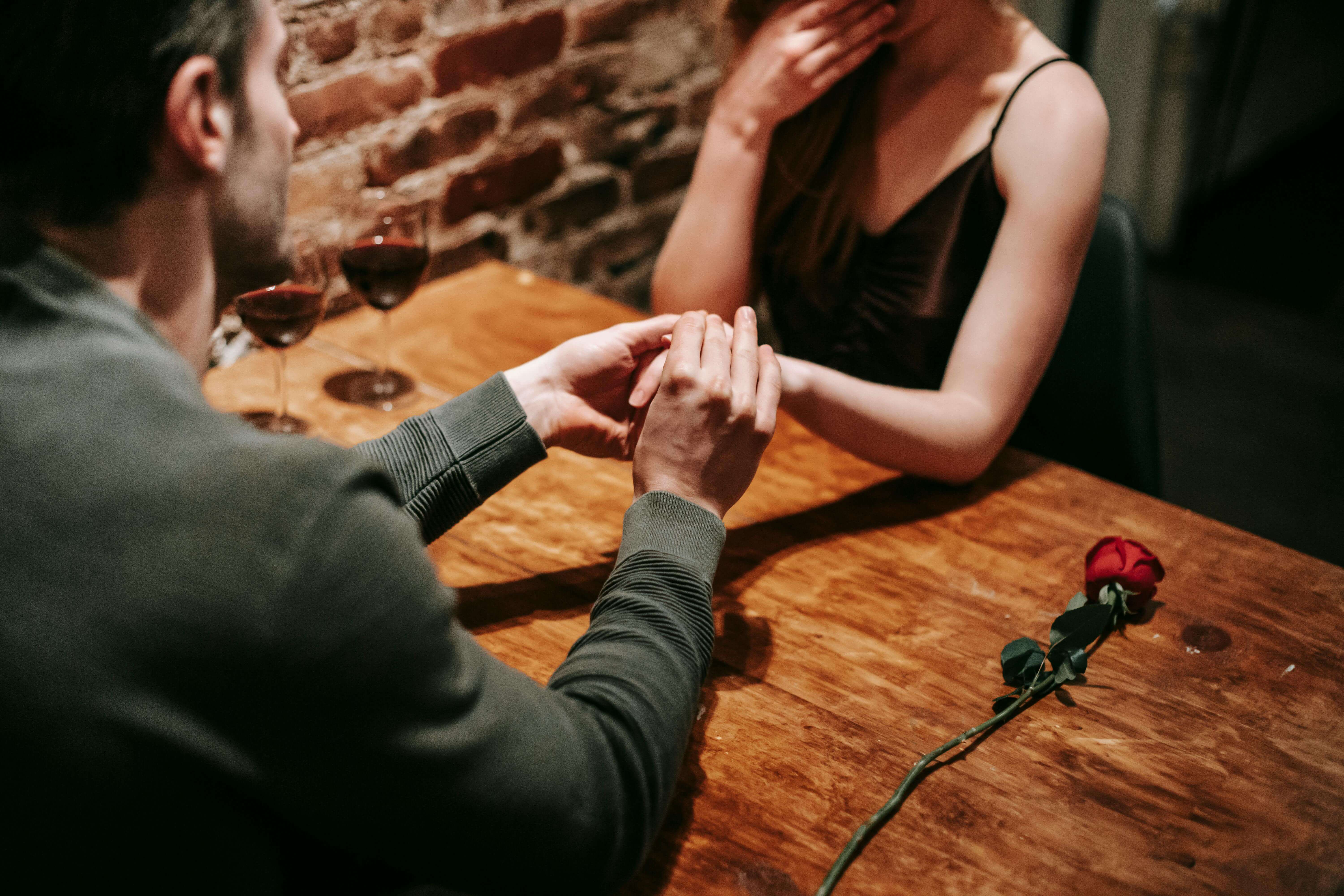 Crop unrecognizable couple in elegant clothes sitting in cafeteria at table with wine in glasses and rose near brick wall while having romantic dinner and holding hand while making proposal