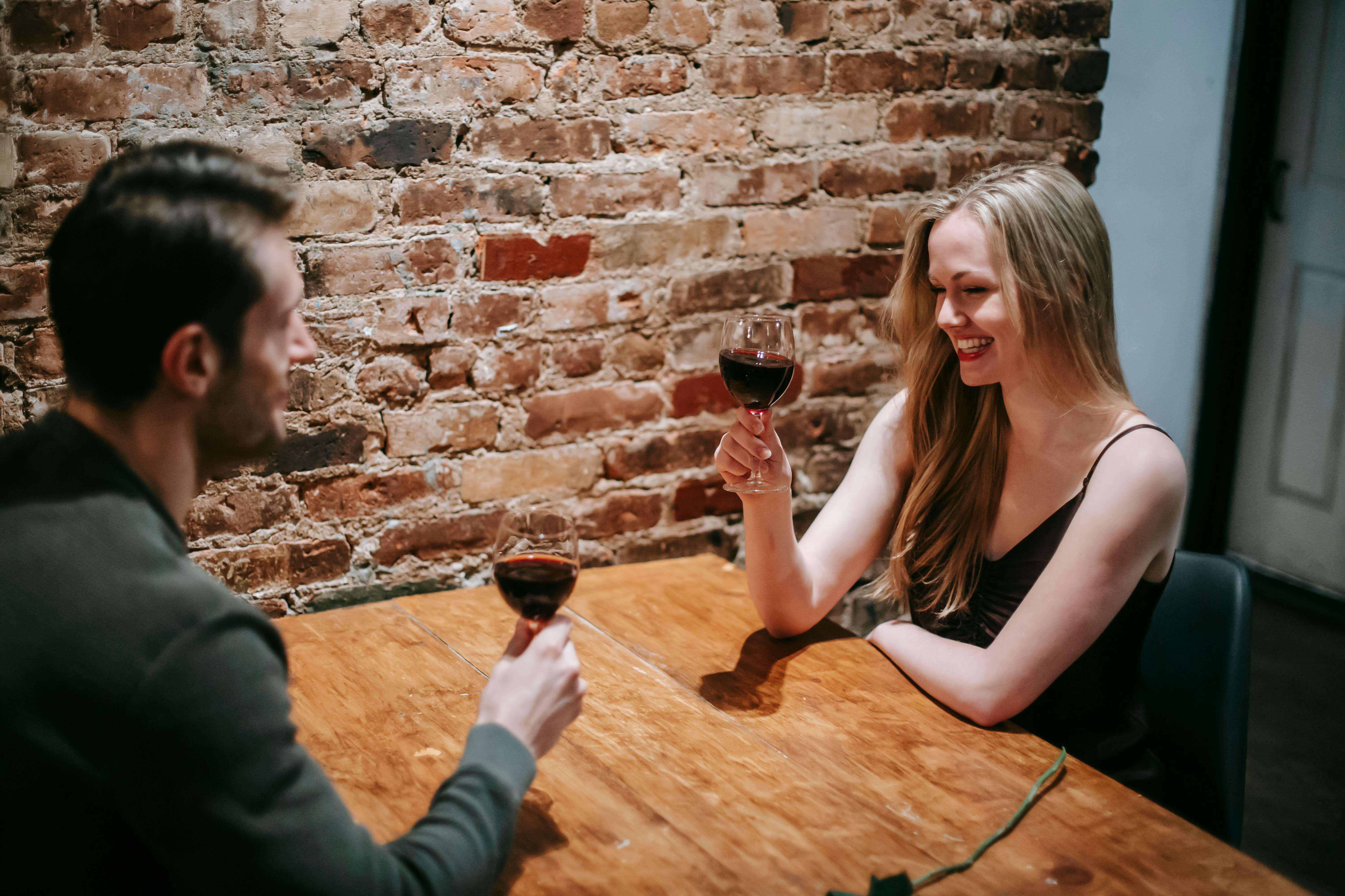 Side view of young smiling couple in elegant clothes sitting in cafeteria at table near brick wall while having romantic dinner with wine in glasses and looking at each other