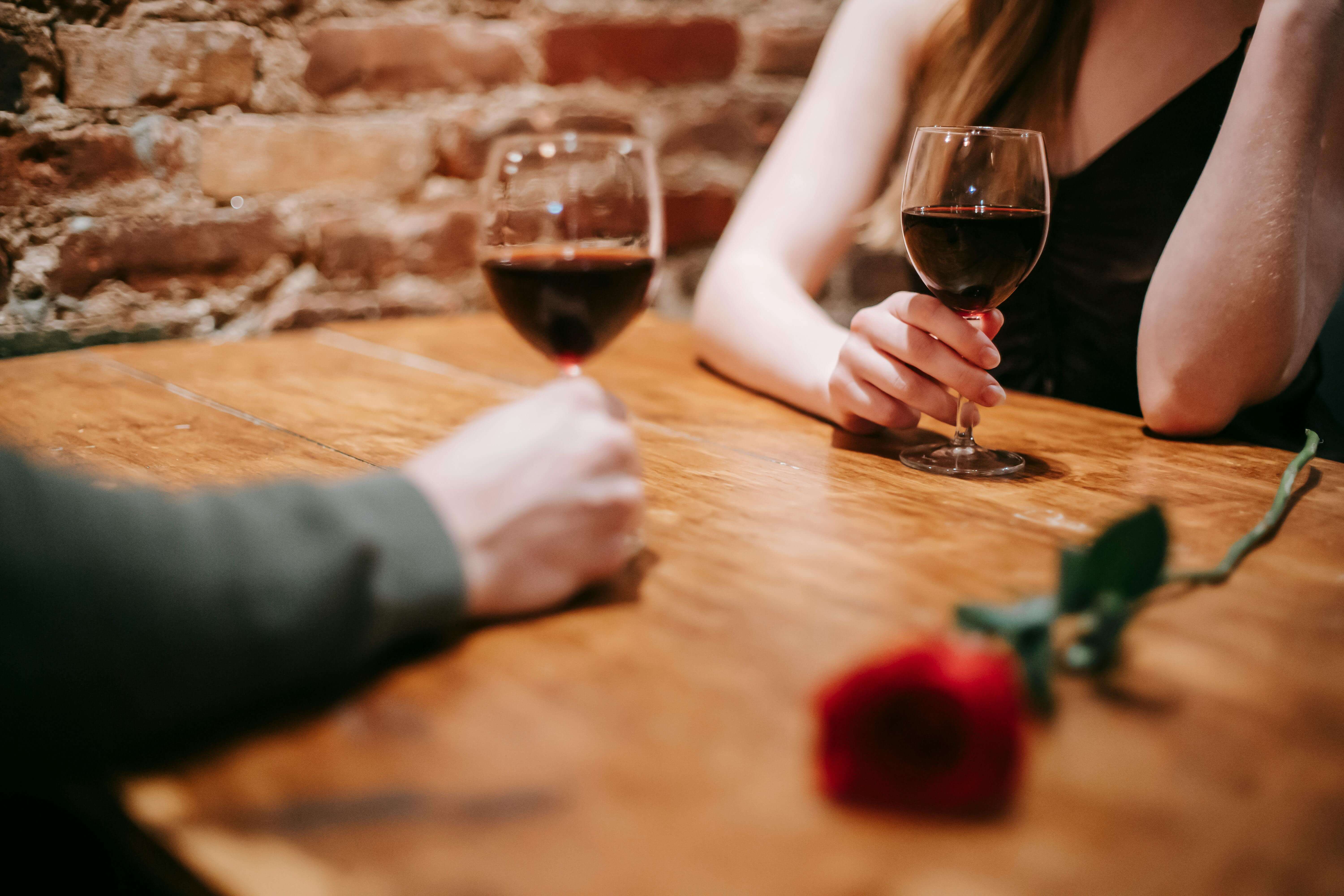 Crop anonymous couple in elegant outfits enjoying romantic date in bar at table with red rose and glasses with wine near brick wall