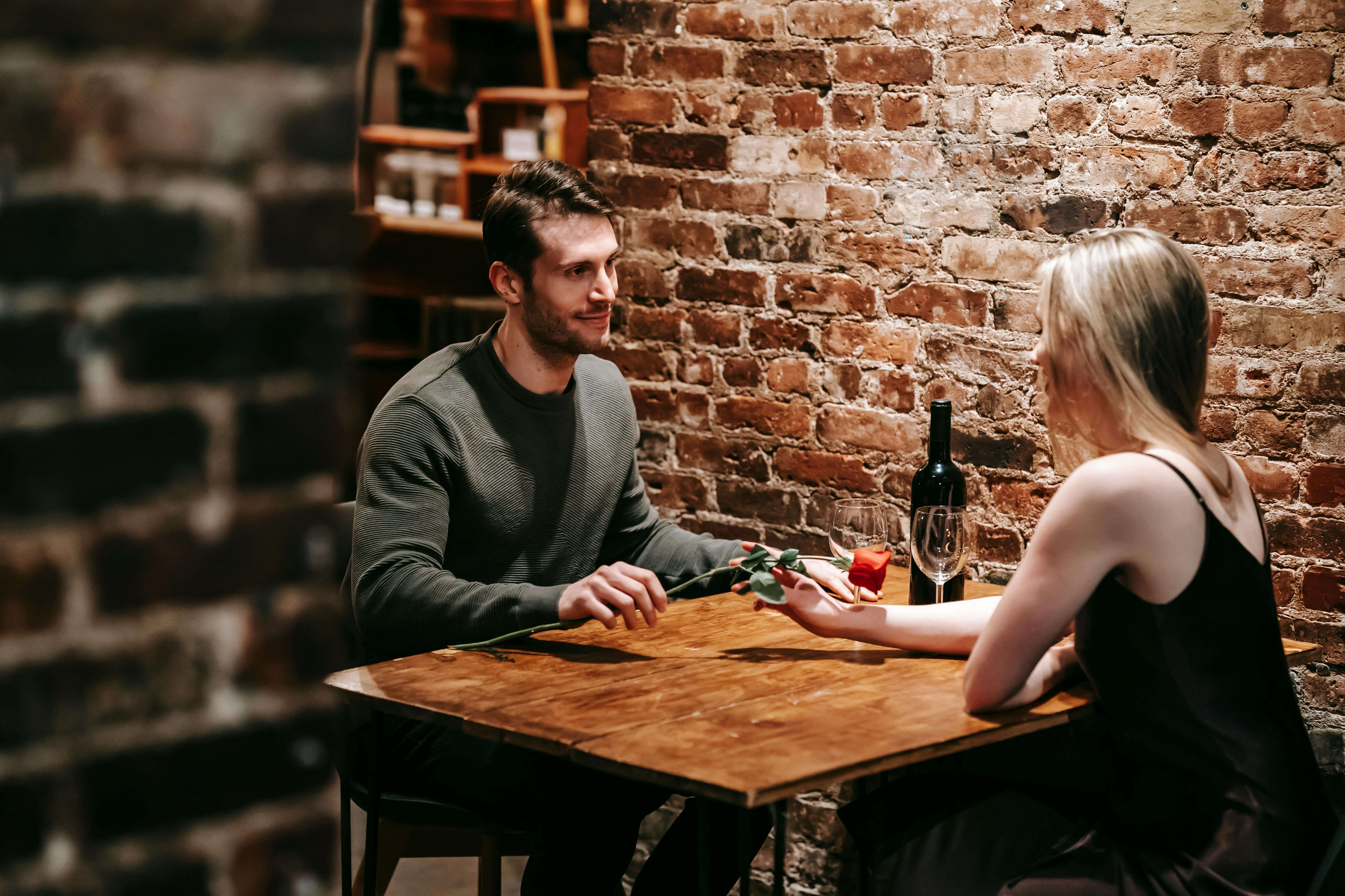 Loving boyfriend giving red rose to girlfriend while sitting near brick wall at wooden table during celebration of event in restaurant