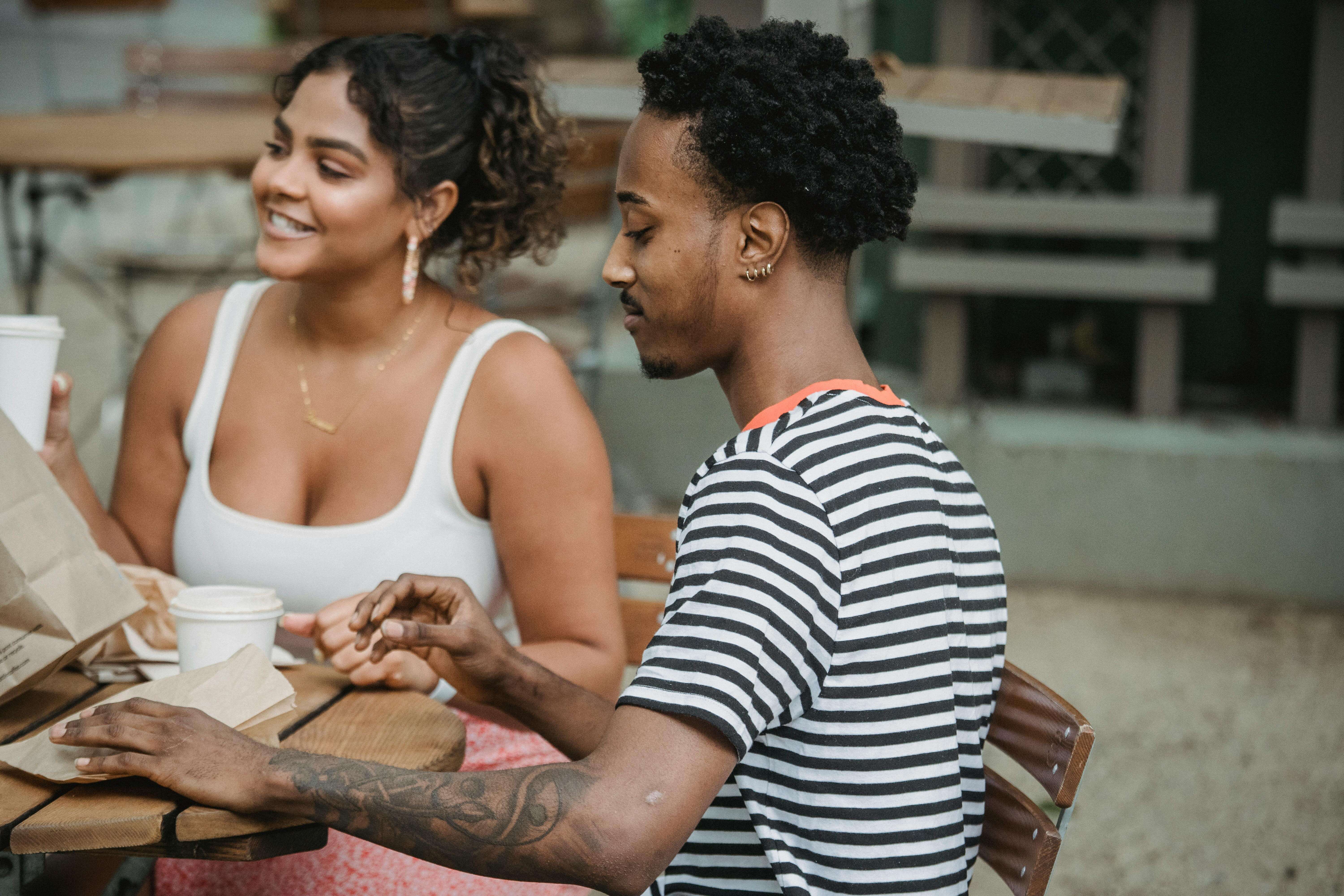 Happy young Hispanic woman and African American man in casual clothes resting on cafe terrace and drinking takeaway coffee during date