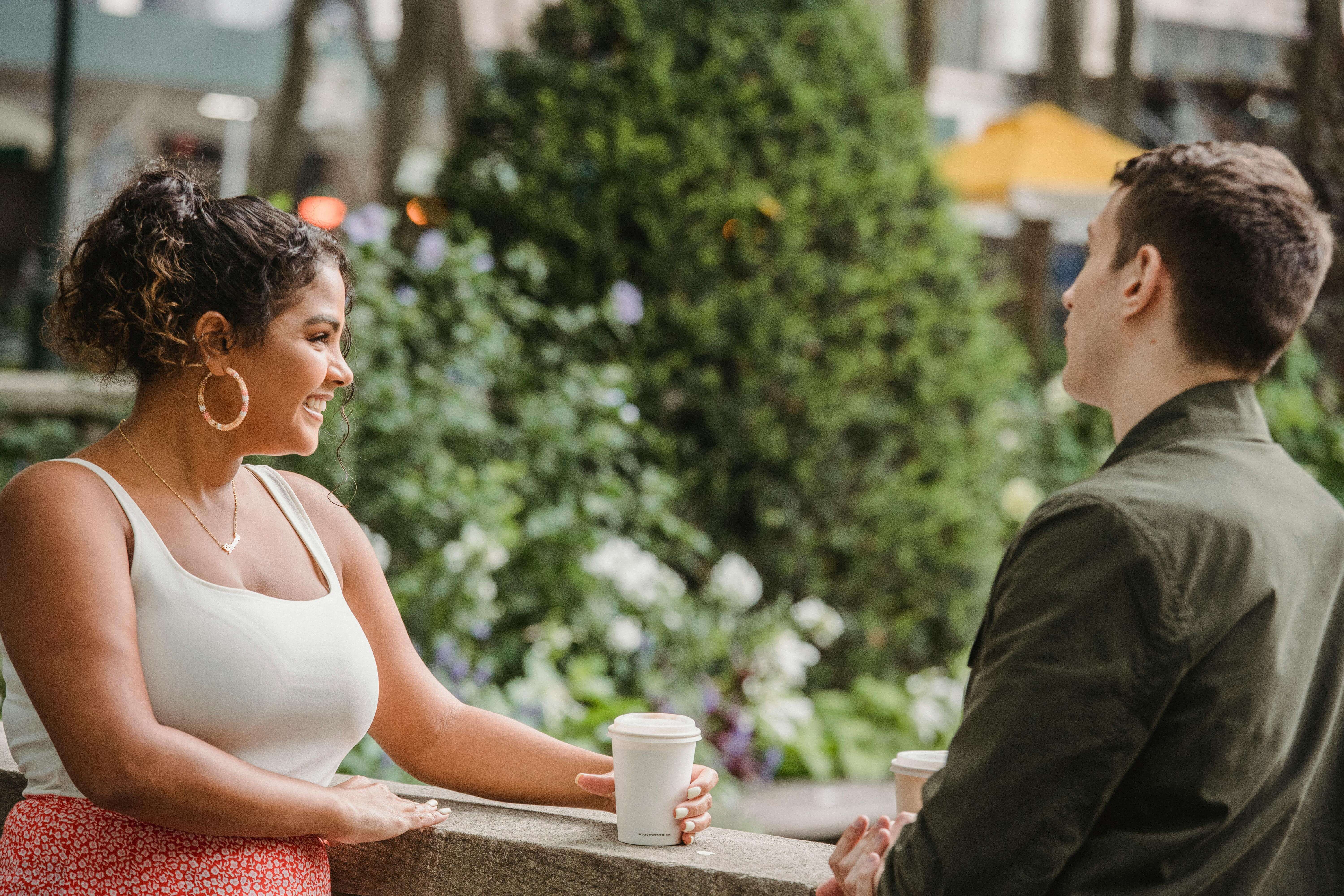 Cheerful young Hispanic woman in casual clothes with boyfriend standing on terrace and smiling while having coffee break together in outdoor cafe