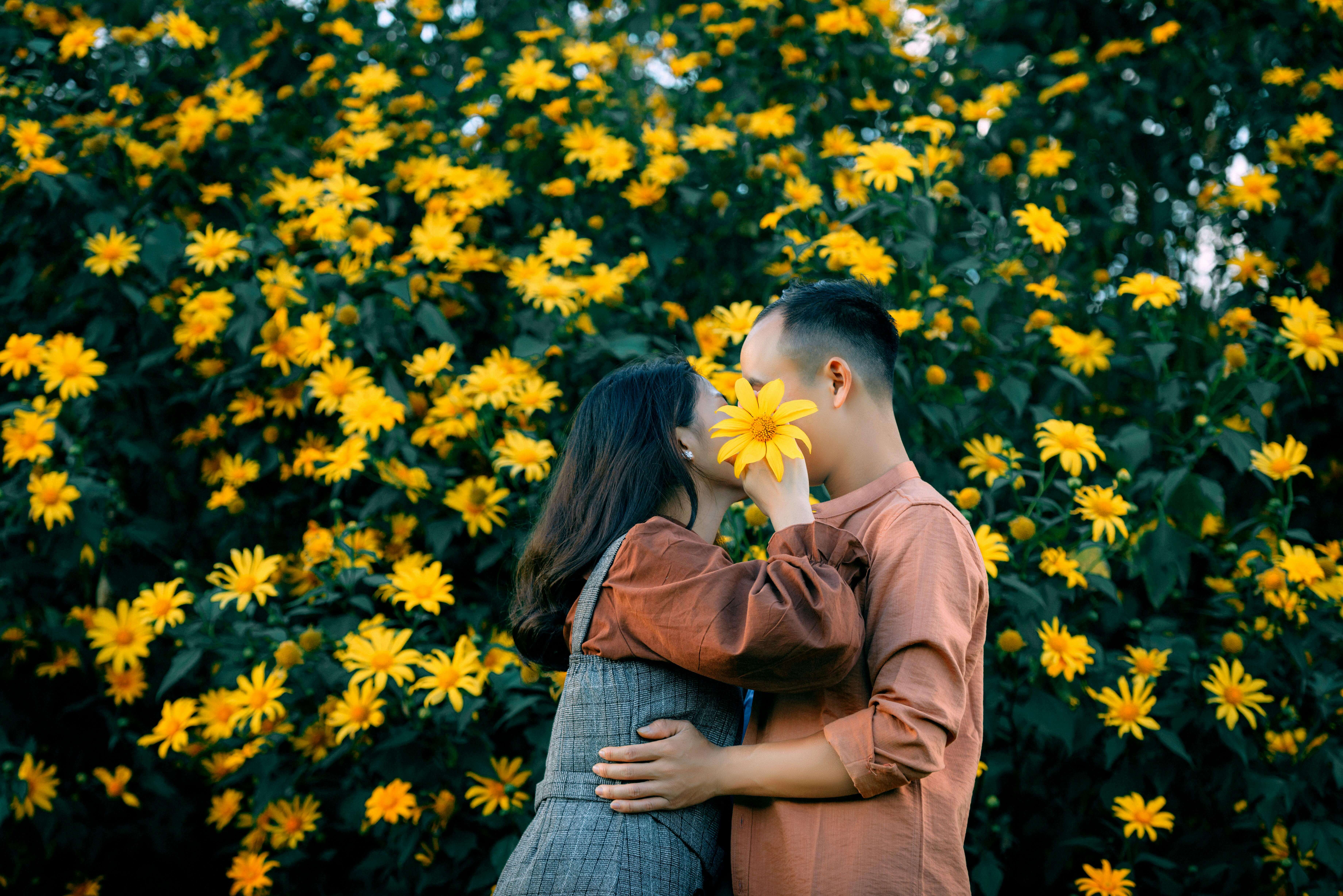 Side view of unrecognizable romantic couple embracing and covering faces with yellow flower while standing near lush blossoming bush in garden