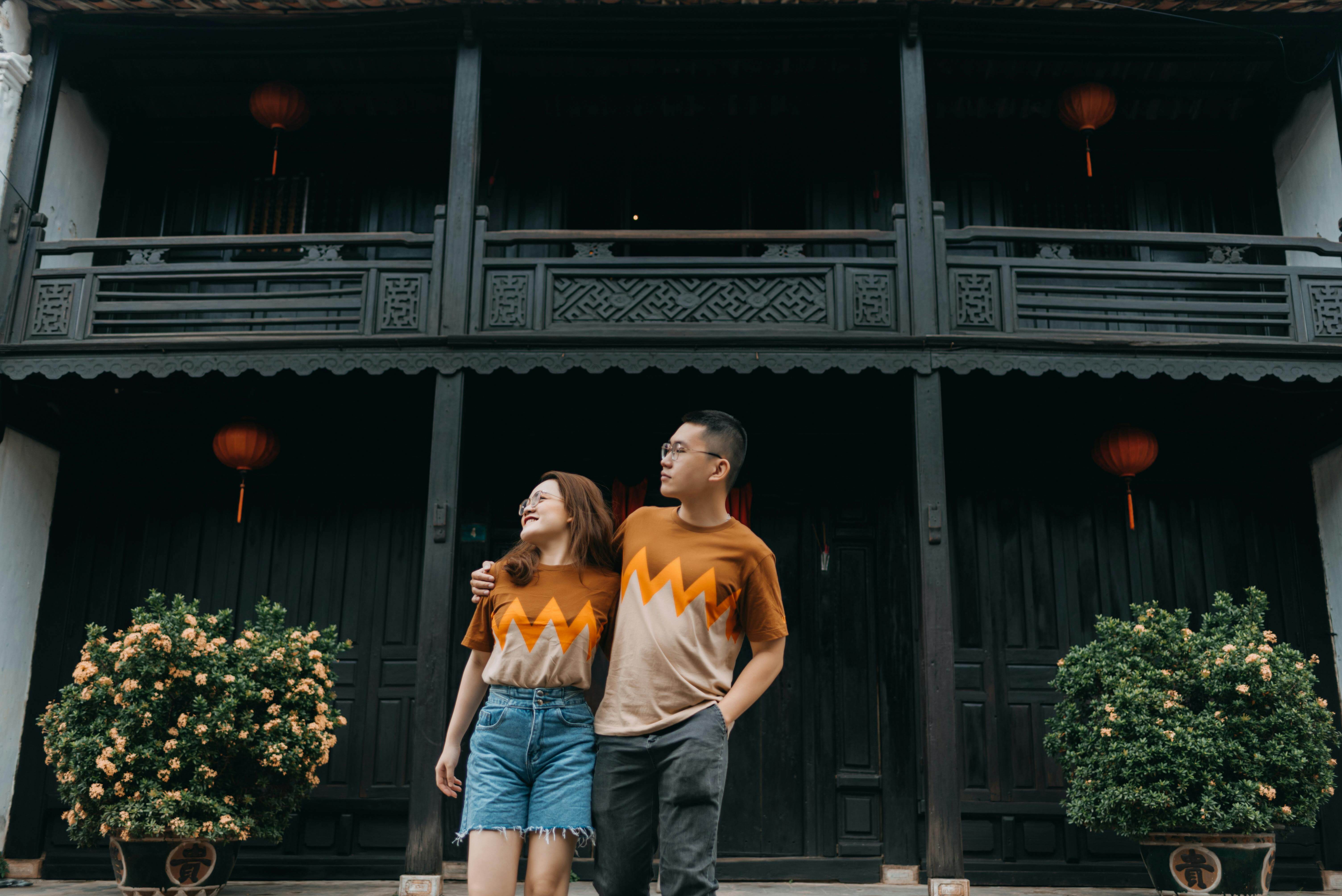 A young Asian couple embracing and looking away, standing outside a traditional building with potted plants and lanterns.