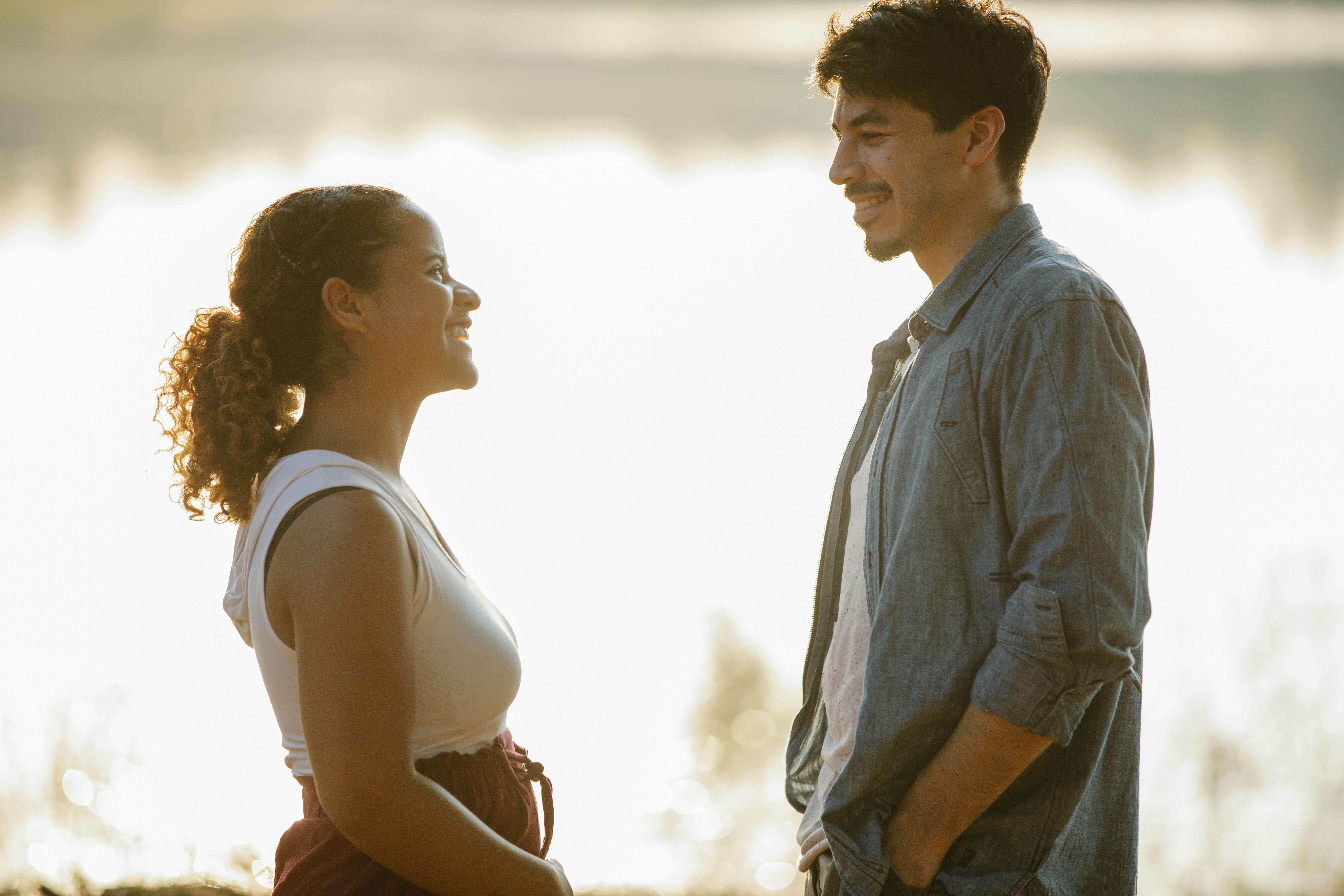 Side view of cheerful young man and Hispanic woman in casual clothes smiling and chatting while relaxing on shore of calm lake during romantic date in nature at sundown