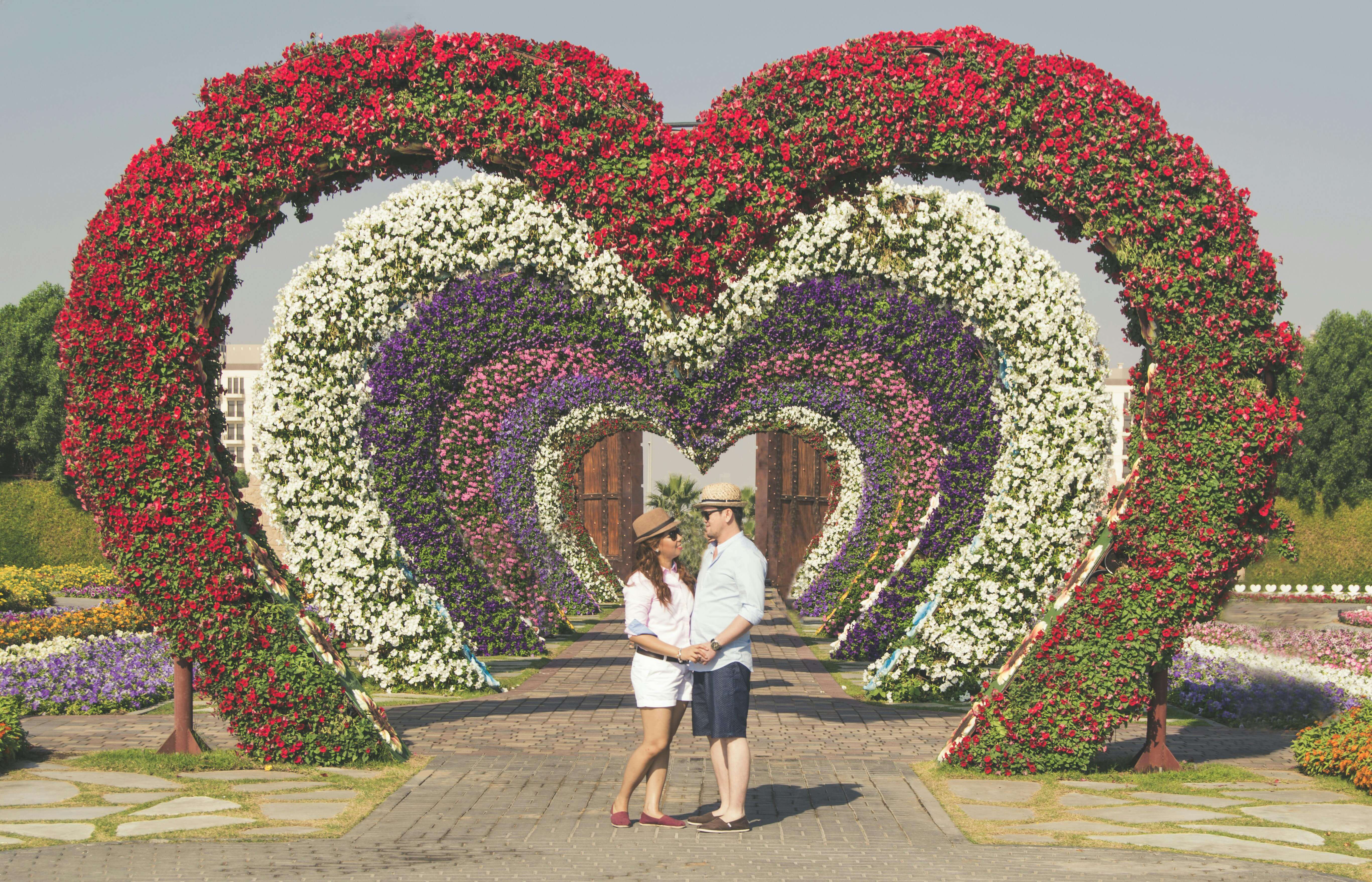 A couple embracing in a vibrant flower garden in Dubai, featuring heart-shaped floral arches.