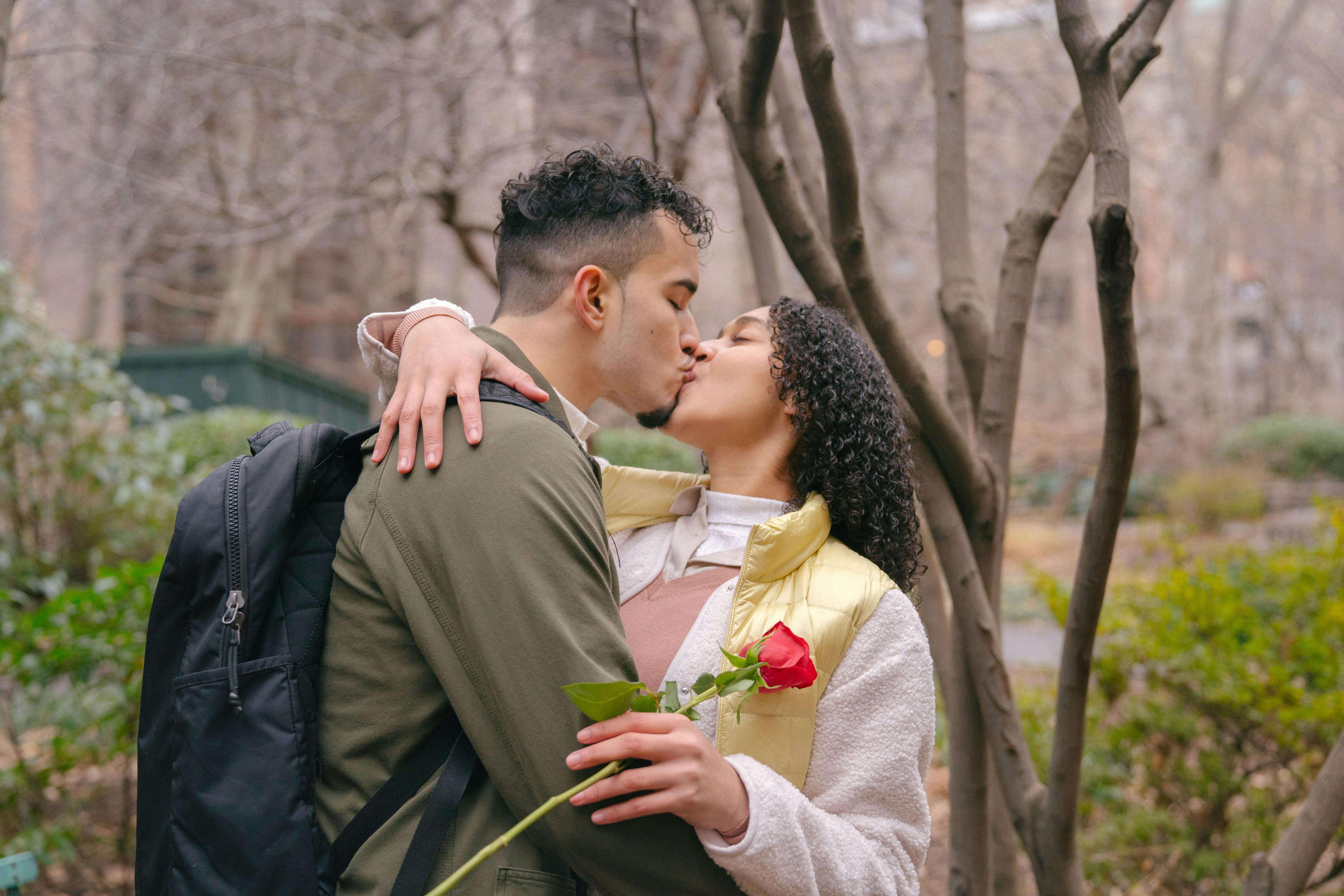 Young Hispanic couple in casual clothes standing in park while hugging and kissing tenderly with red rose in hand in daytime near trees and green bushes
