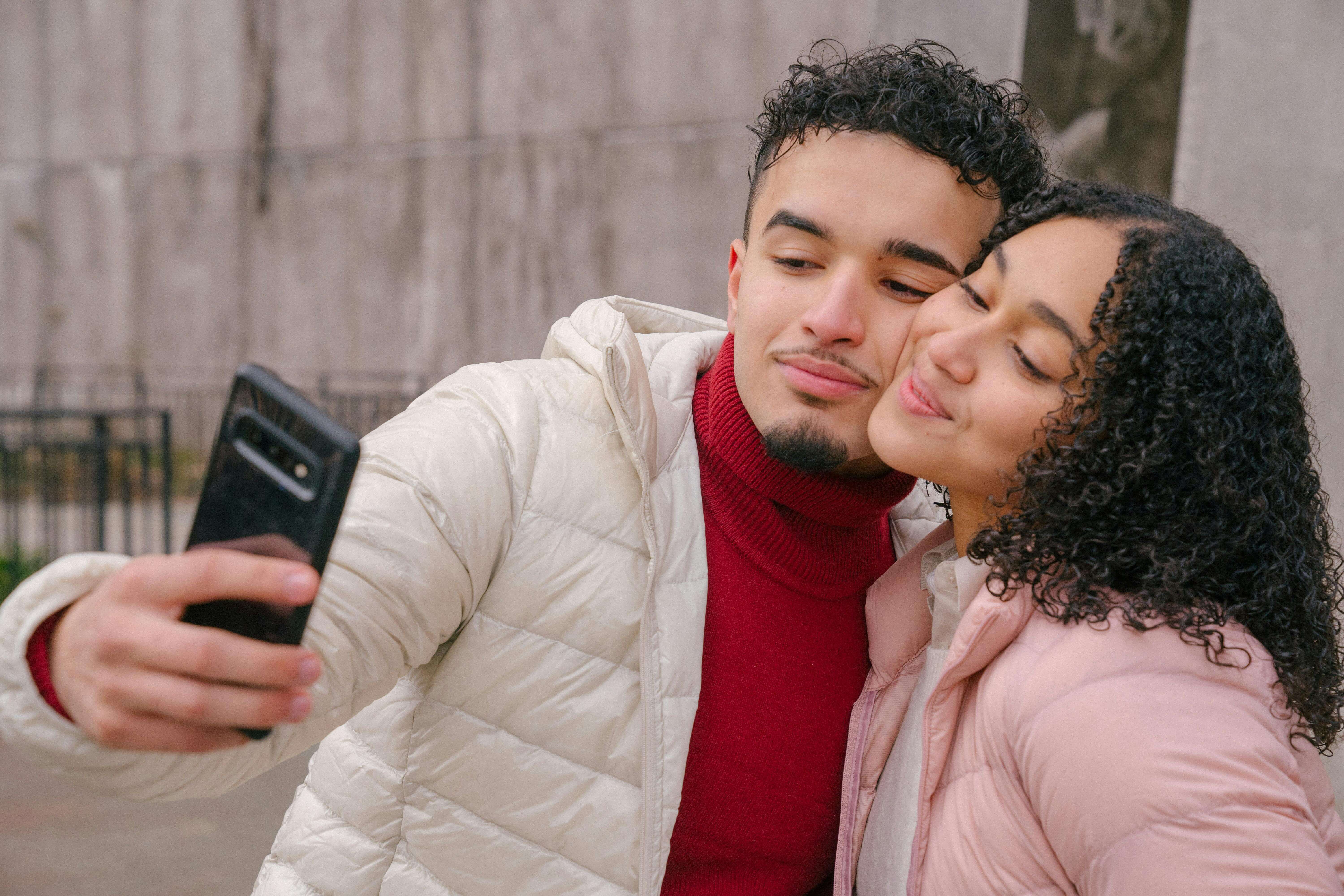 Enamored young Hispanic couple in warm casual clothes smiling and touching cheeks while taking selfie on mobile phone standing on street