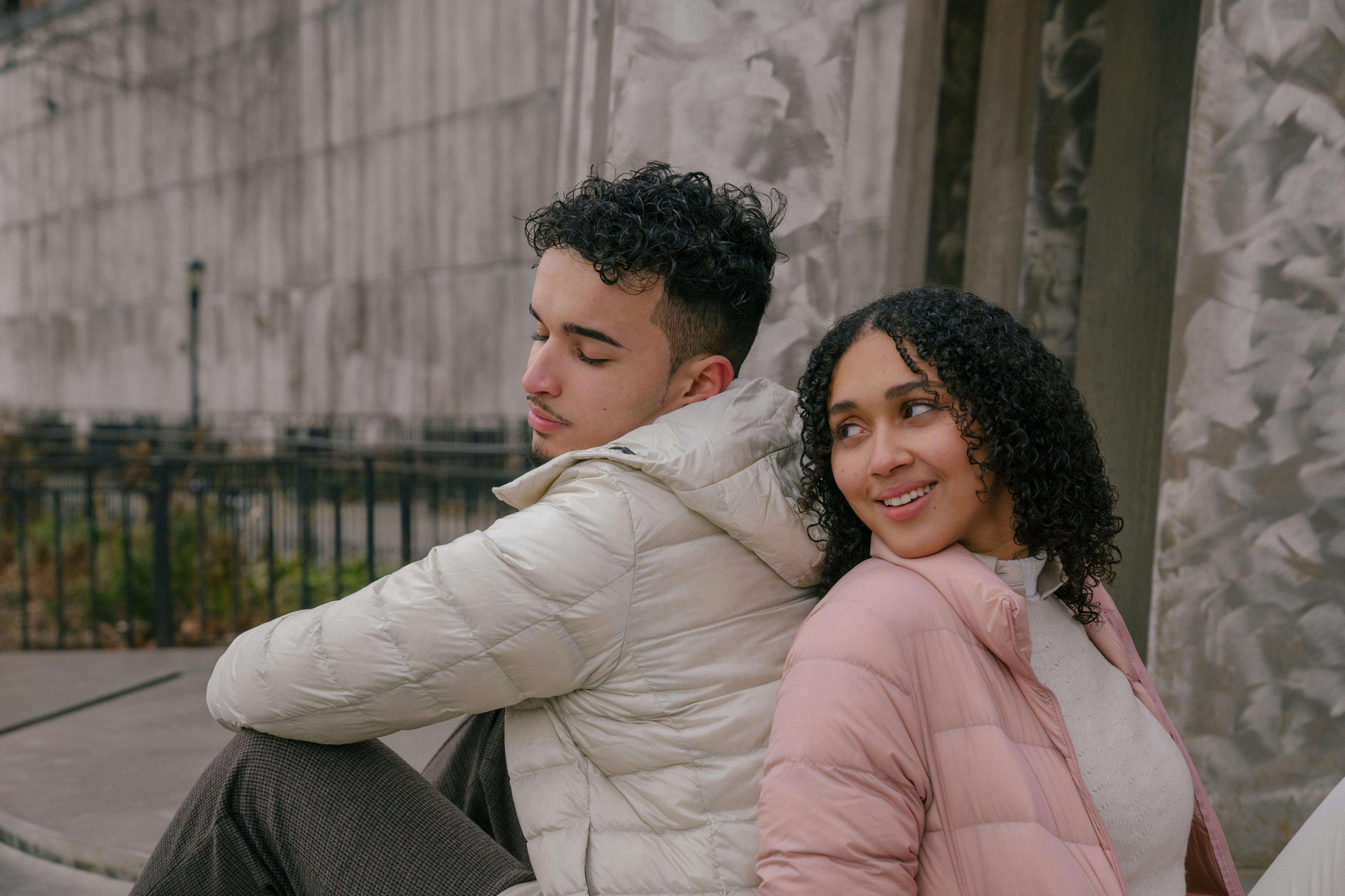 Relaxed happy young ethnic man and woman with curly hair in casual clothes sitting back to back during date in park