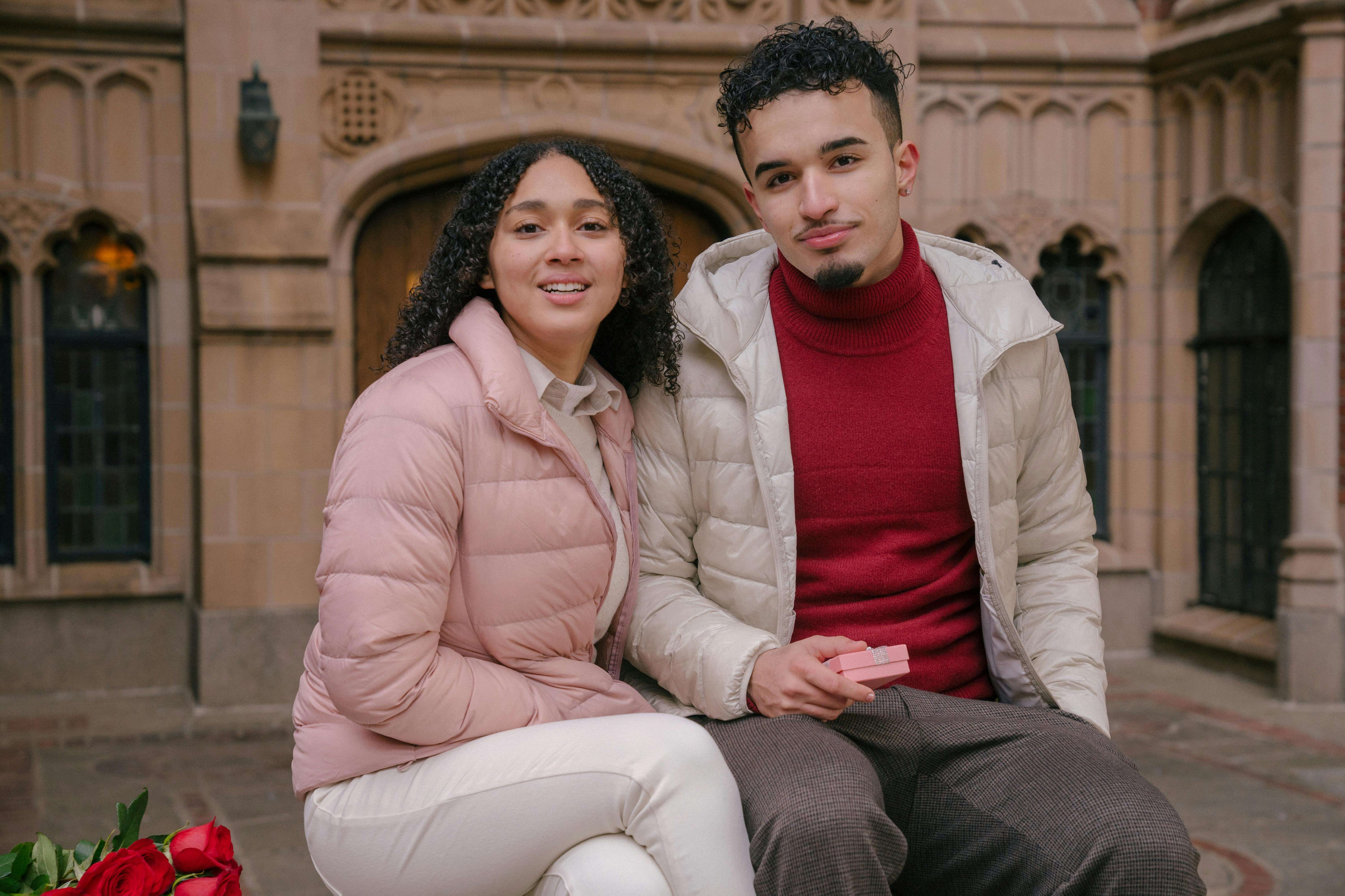 Content young Hispanic couple in stylish outfits with small gift box and bunch of red roses sitting on bench near historic building and looking at camera