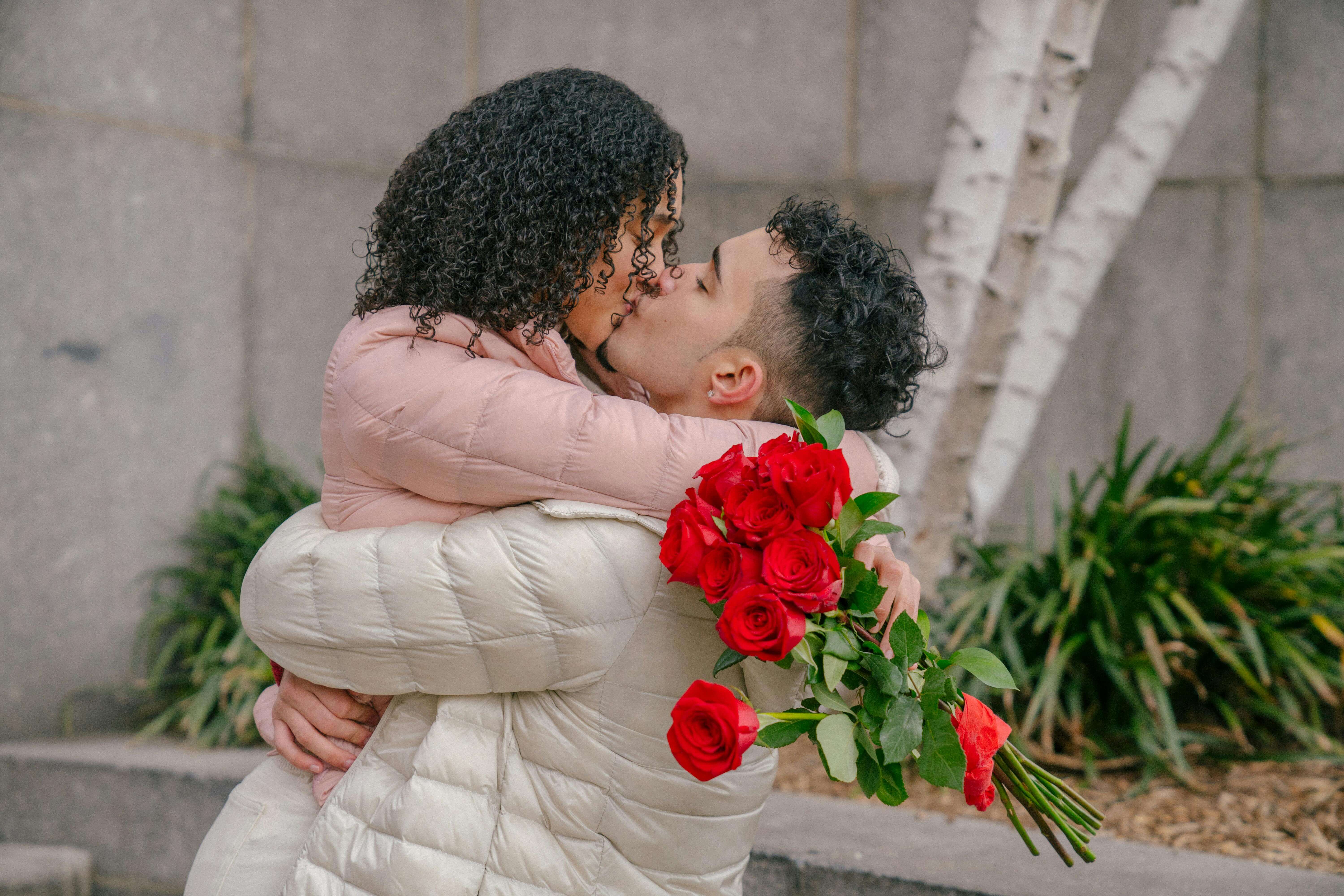A young couple shares a romantic embrace outdoors, holding a bouquet of red roses.