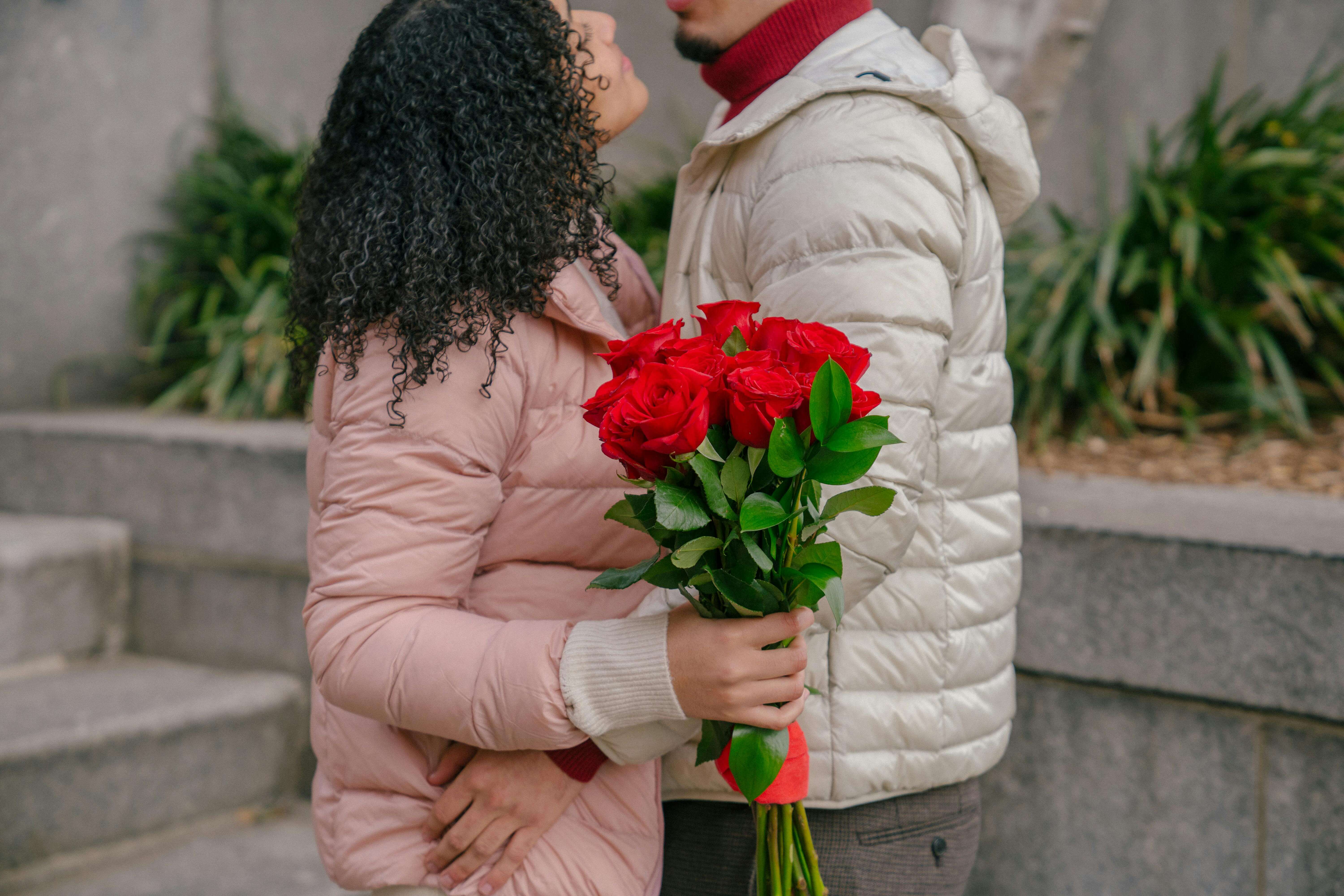 Side view of crop couple wearing warm clothes standing close with bunch of tied roses and embracing