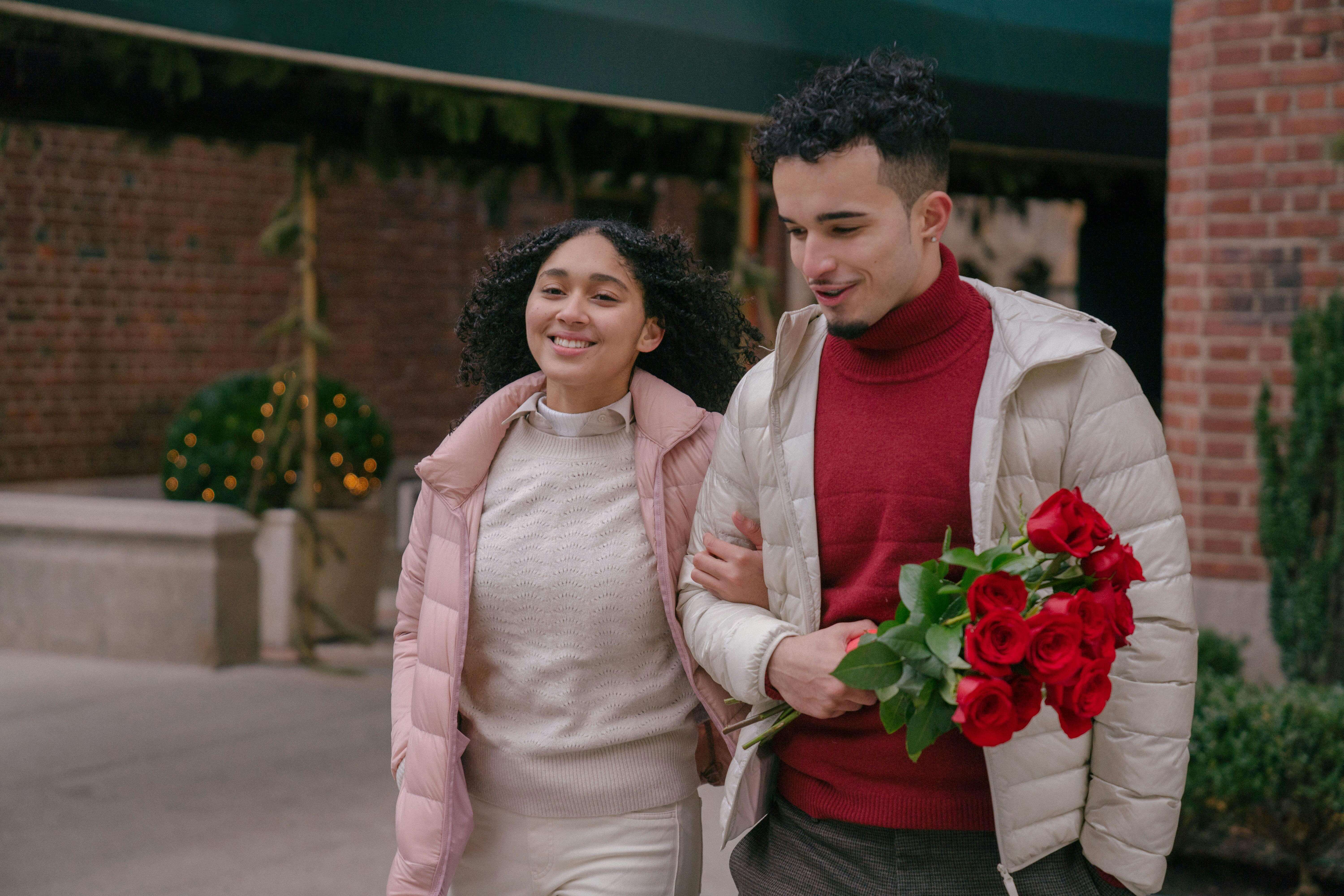 A happy couple strolls arm in arm outside, holding a bouquet of red roses.
