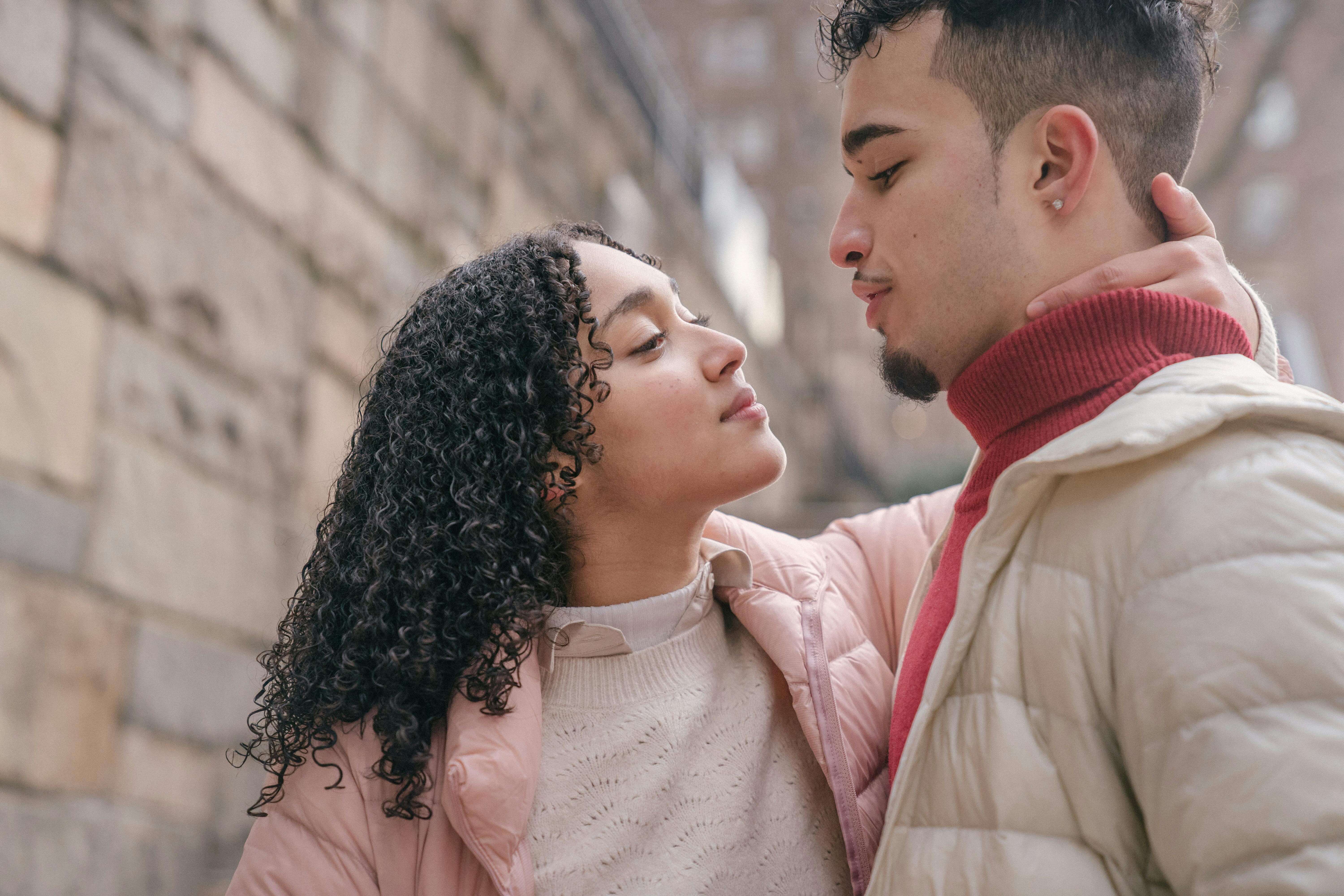 Hispanic couple sharing an affectionate moment outdoors by a stone wall in a city setting.