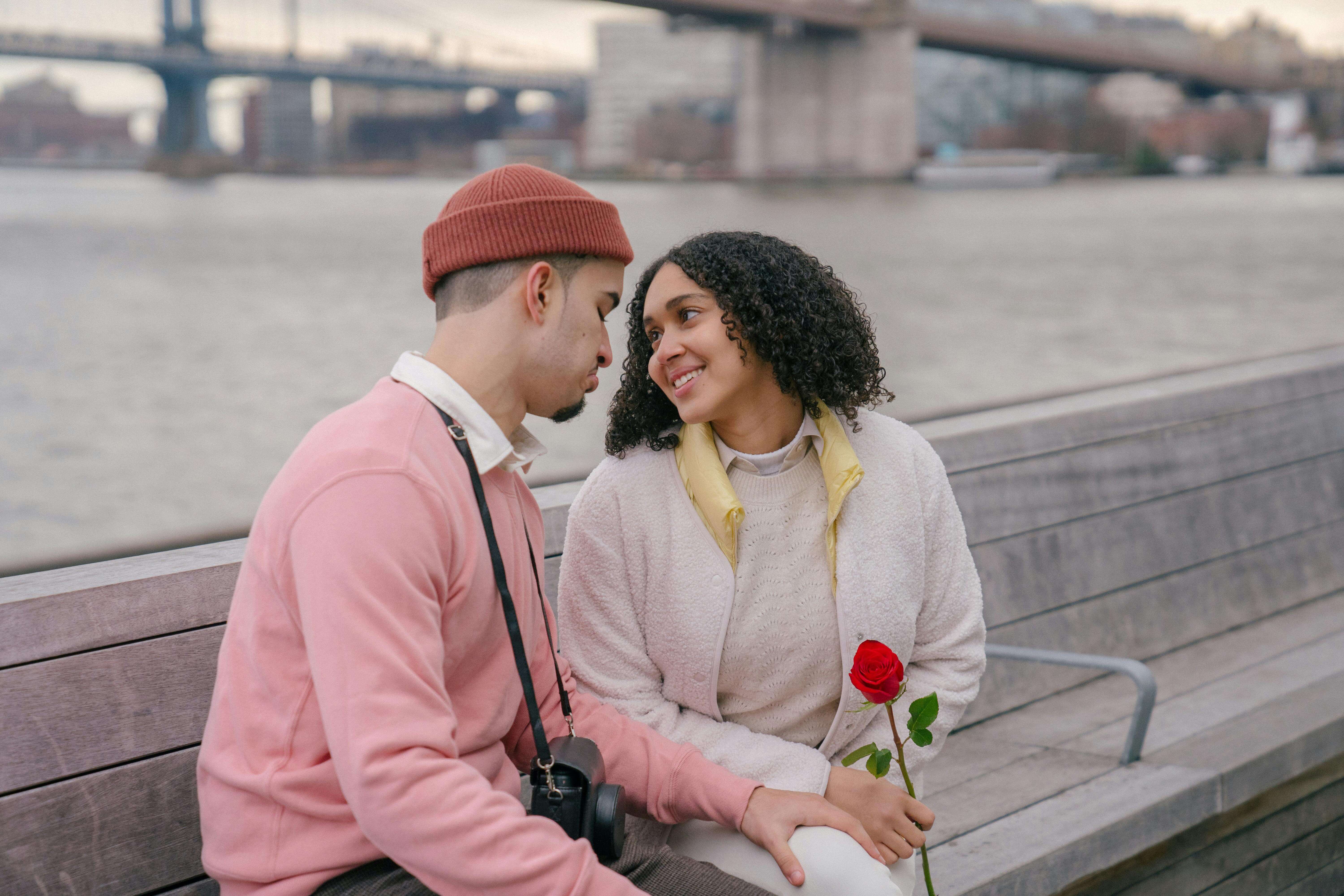 Cheerful Hispanic couple with red rose sitting on wooden bench on seafront near calm water on street during romantic date