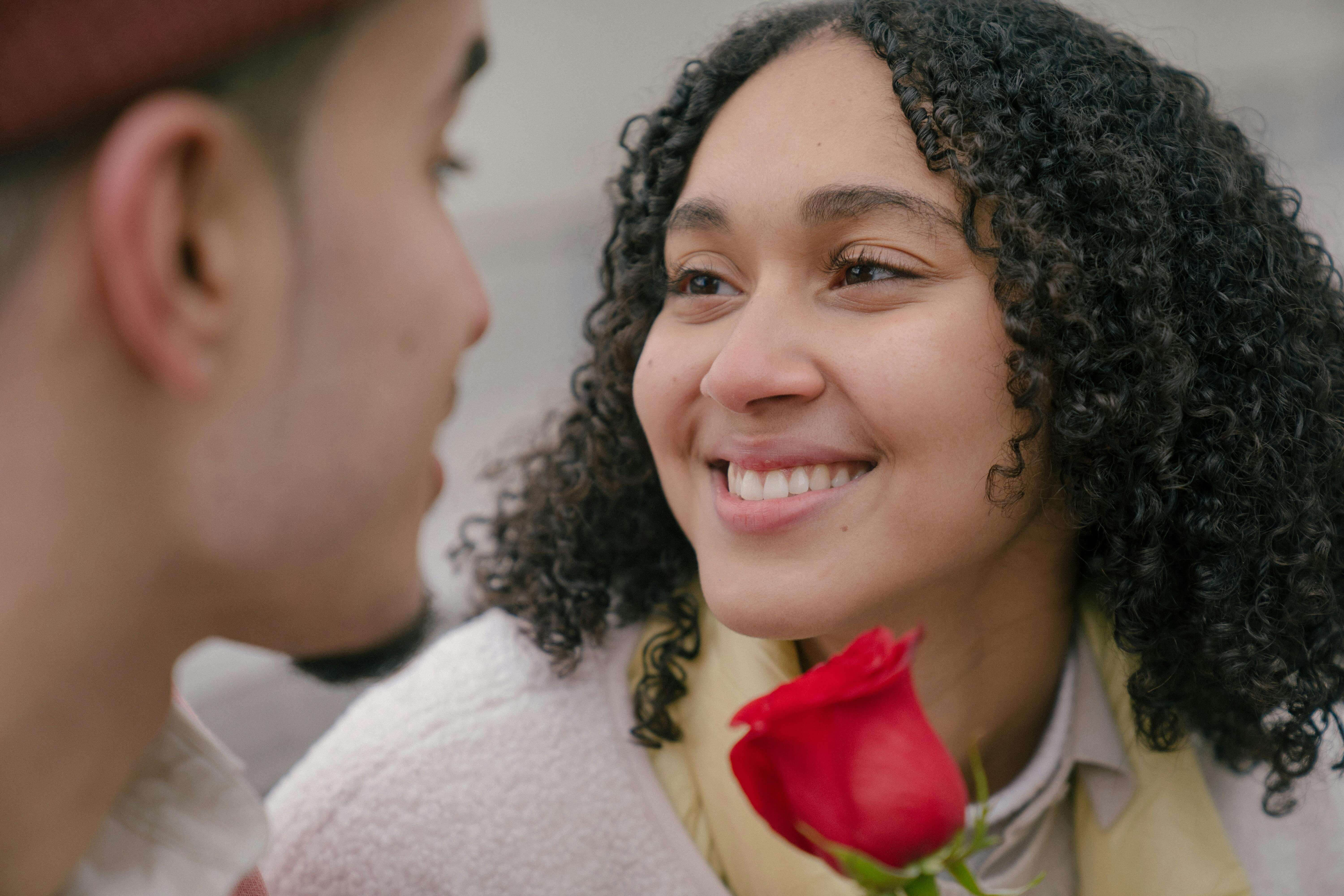 A cheerful young couple sharing a romantic moment outdoors, smiling fondly with a red rose between them.