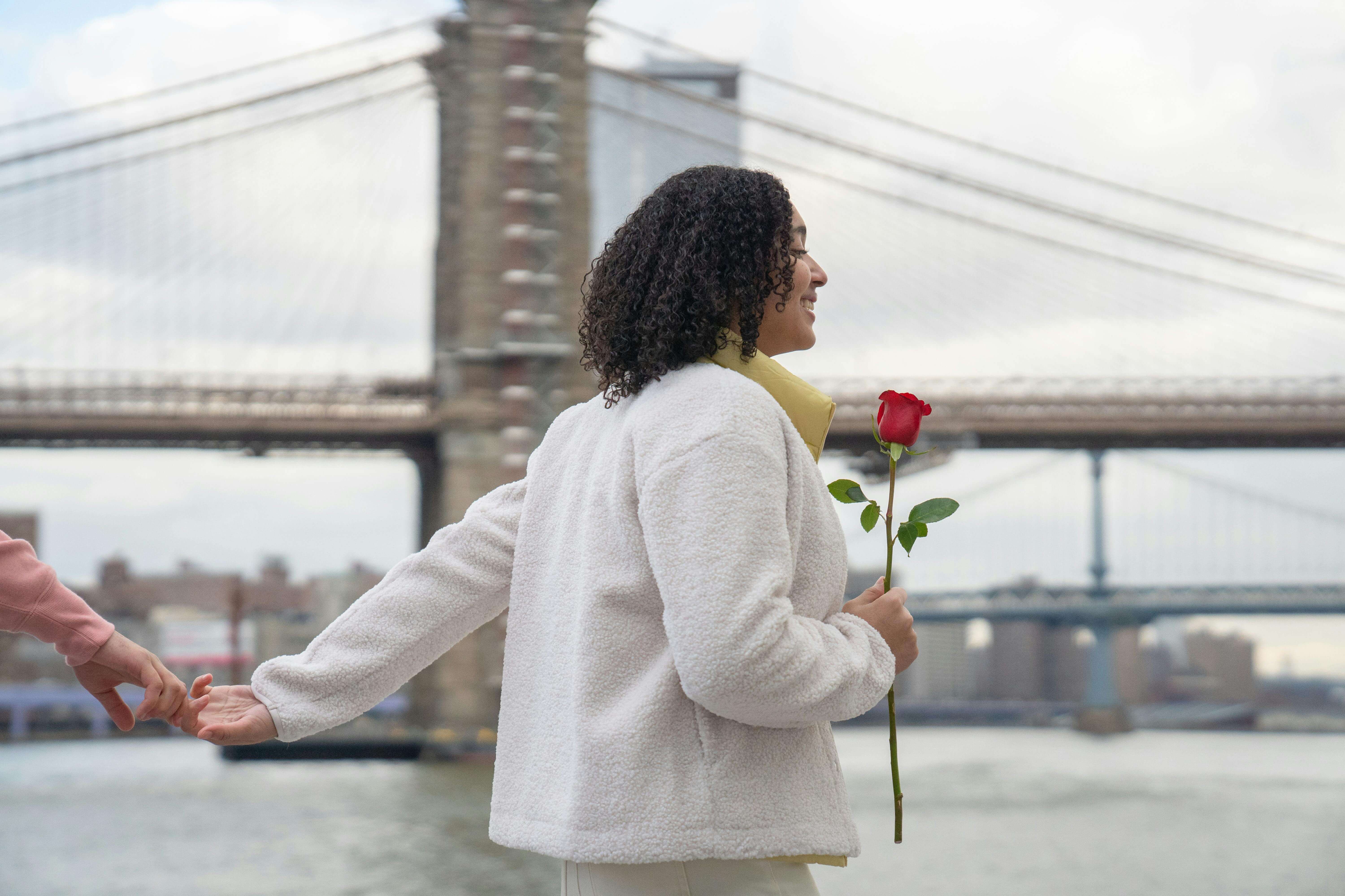 Cheerful Hispanic female with red flower holding hands with faceless boyfriend while strolling on embankment near sea against bridge during date