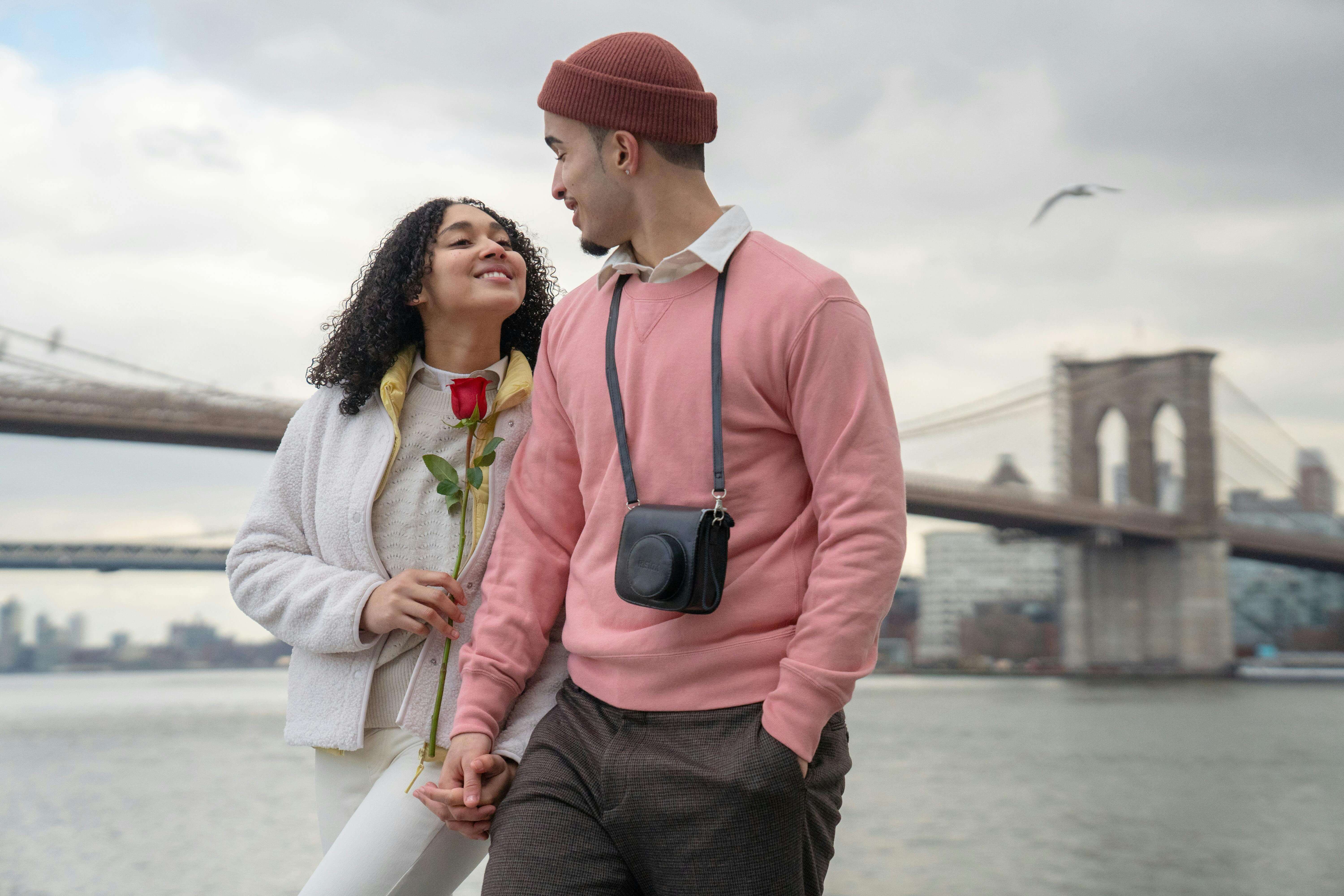 A happy couple holds a rose while strolling by the Brooklyn Bridge waterfront on a cloudy day.