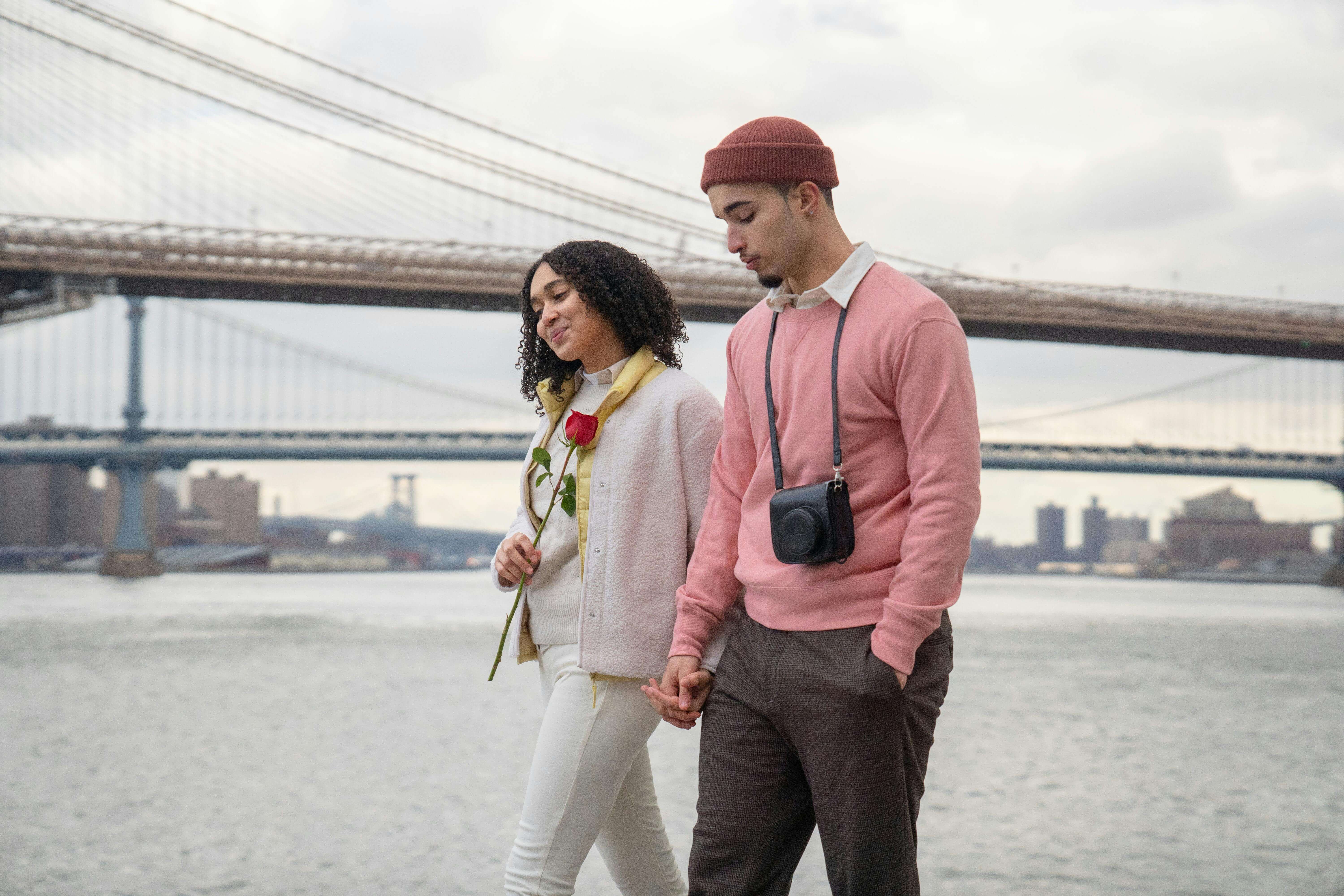A couple walks hand in hand by the Brooklyn waterfront, embracing a romantic moment.