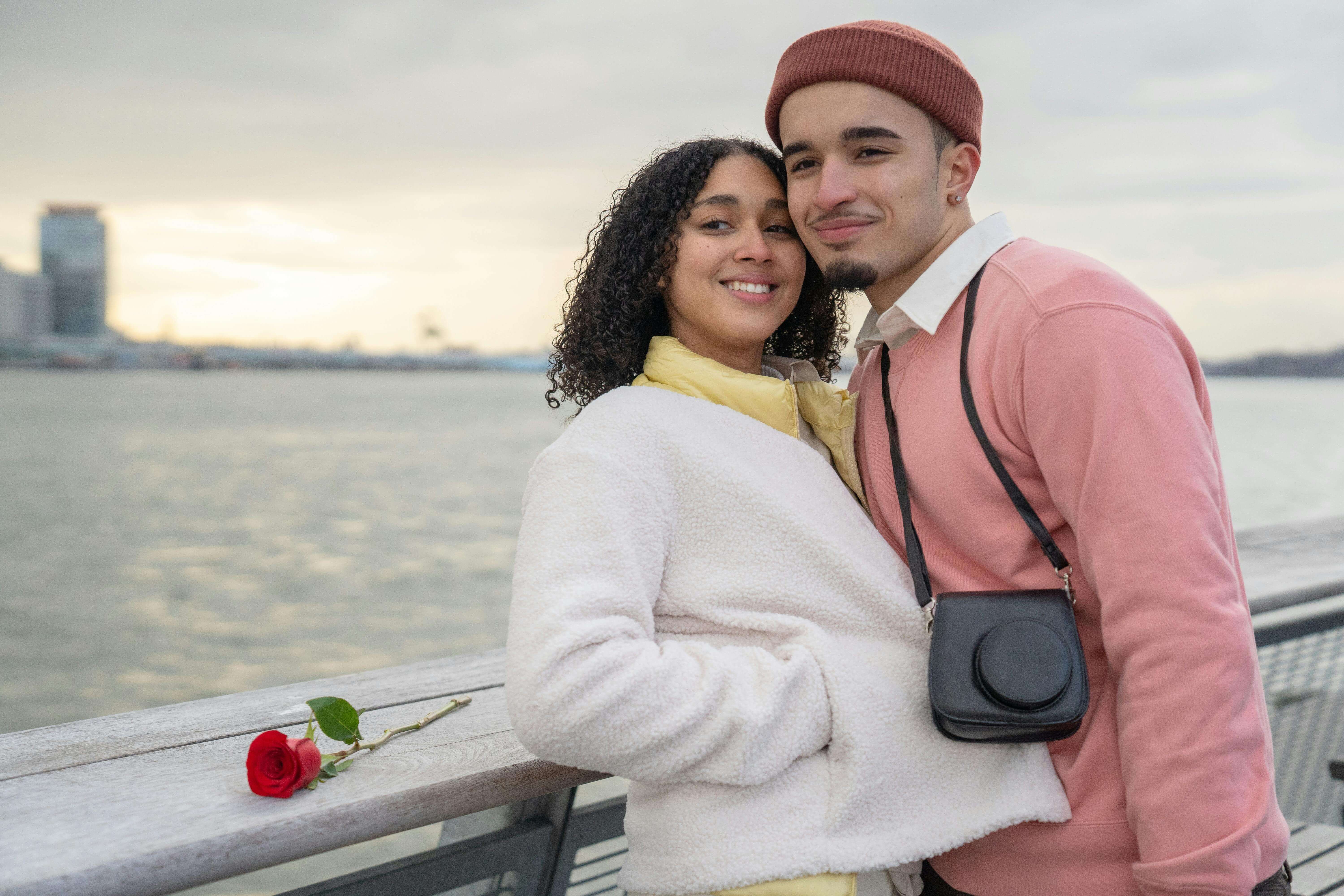 Cheerful Hispanic couple with photo camera looking away while standing near railing on waterfront near sea during romantic date in city